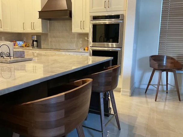 Modern kitchen with white cabinets, a marble island counter, and two wooden barstools, with an oven and a window with blinds.