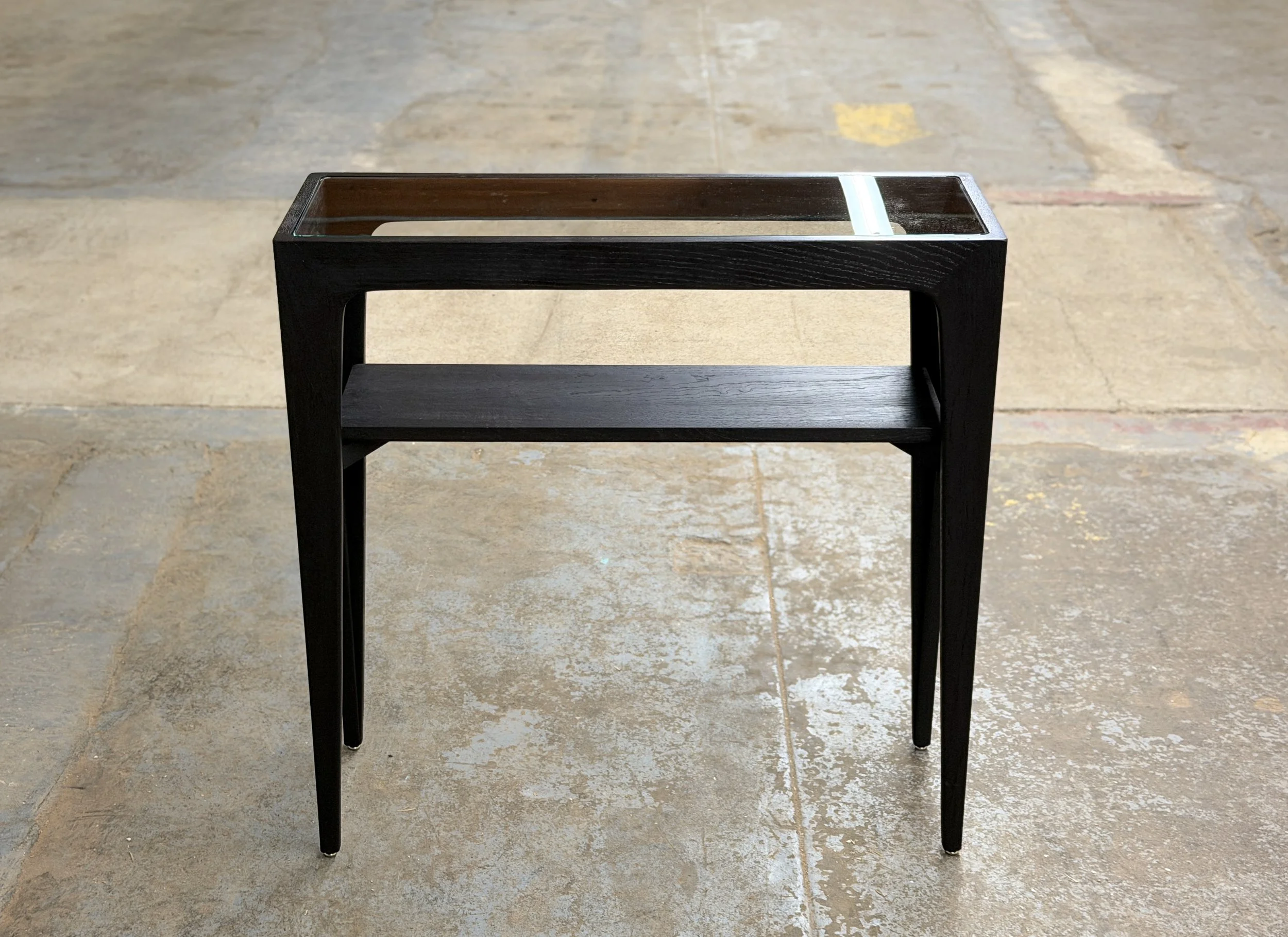 A black wooden side table with a glass top on a concrete floor.