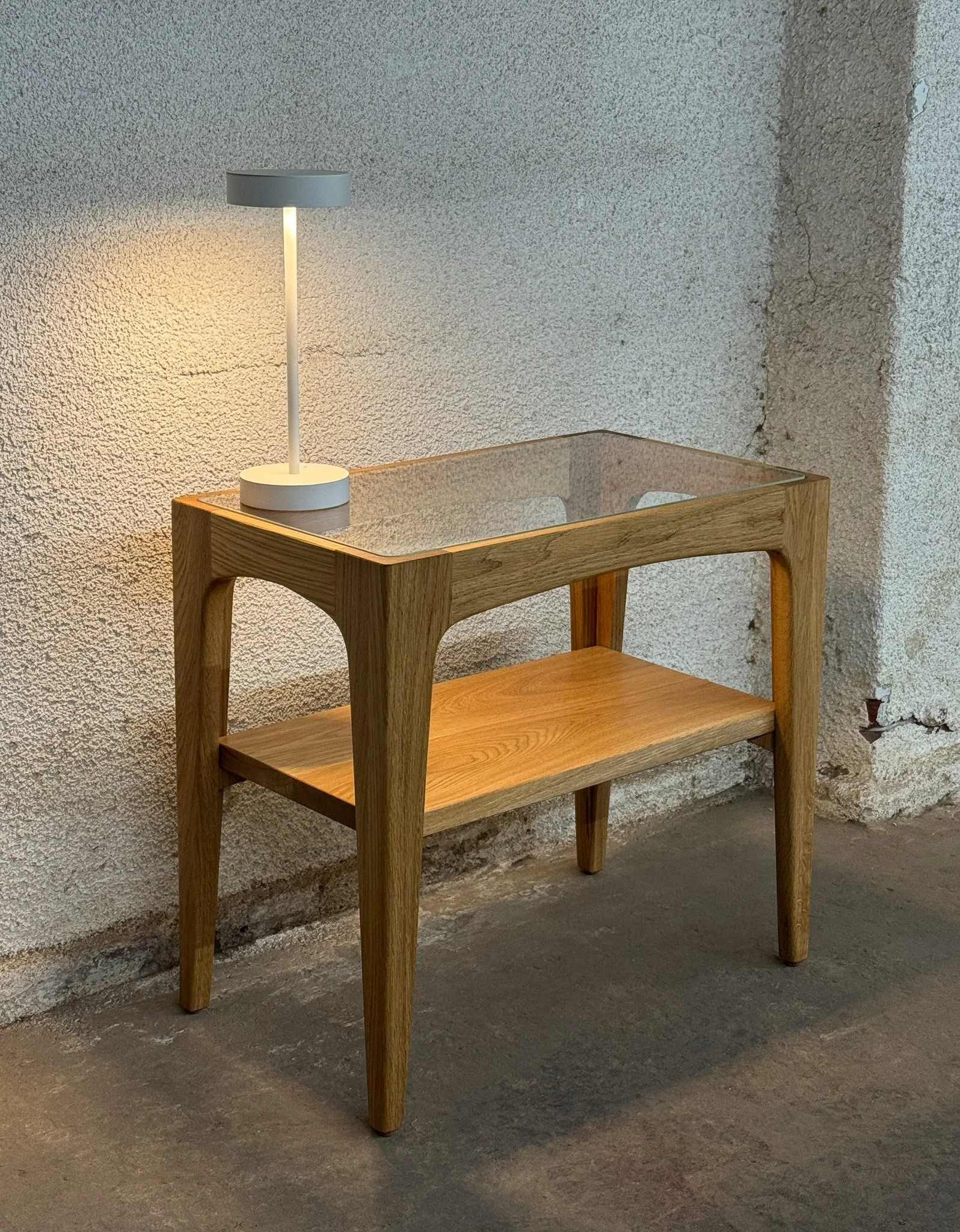 A modern wooden side table with a glass top and a lower shelf, next to a small white table lamp with a gray lampshade, against a textured wall.