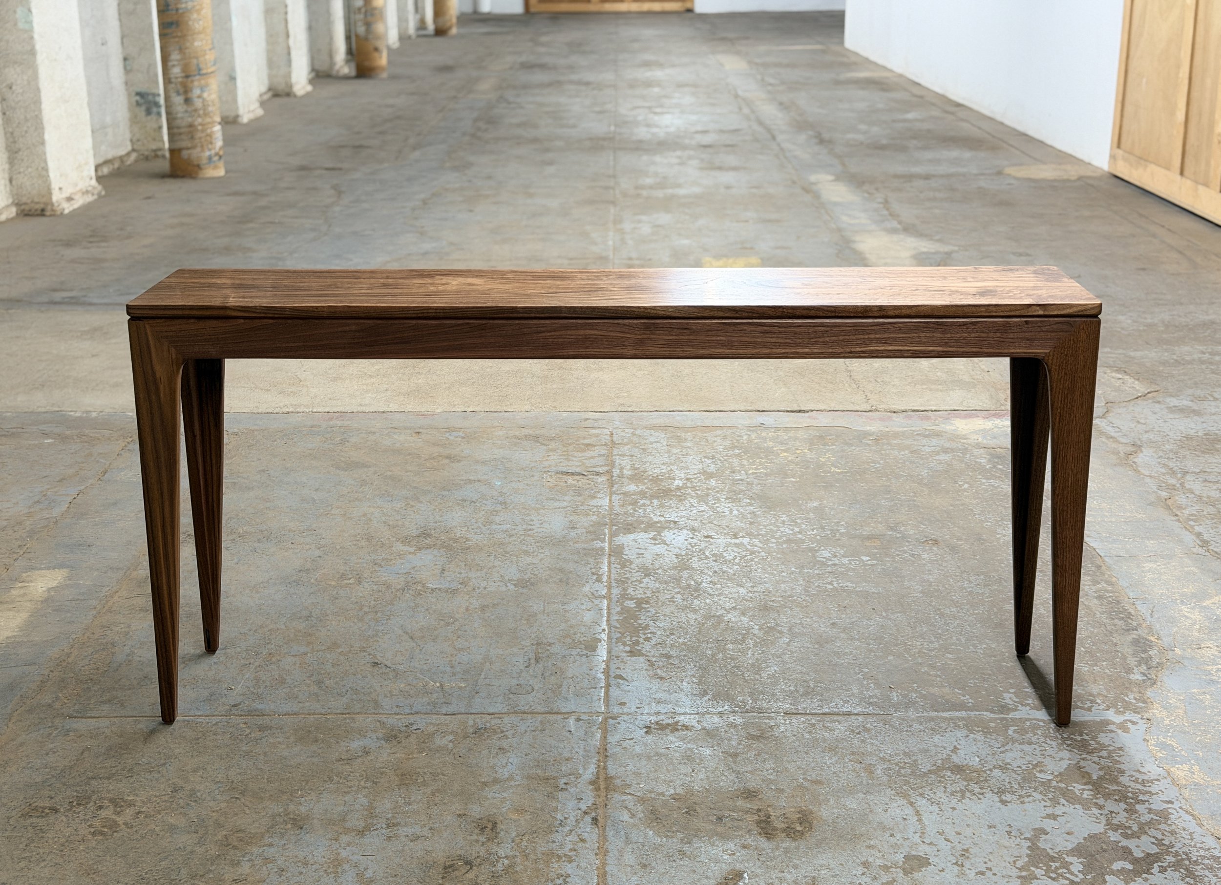 A wooden table with four tapered legs placed on a concrete floor in an unfinished room.