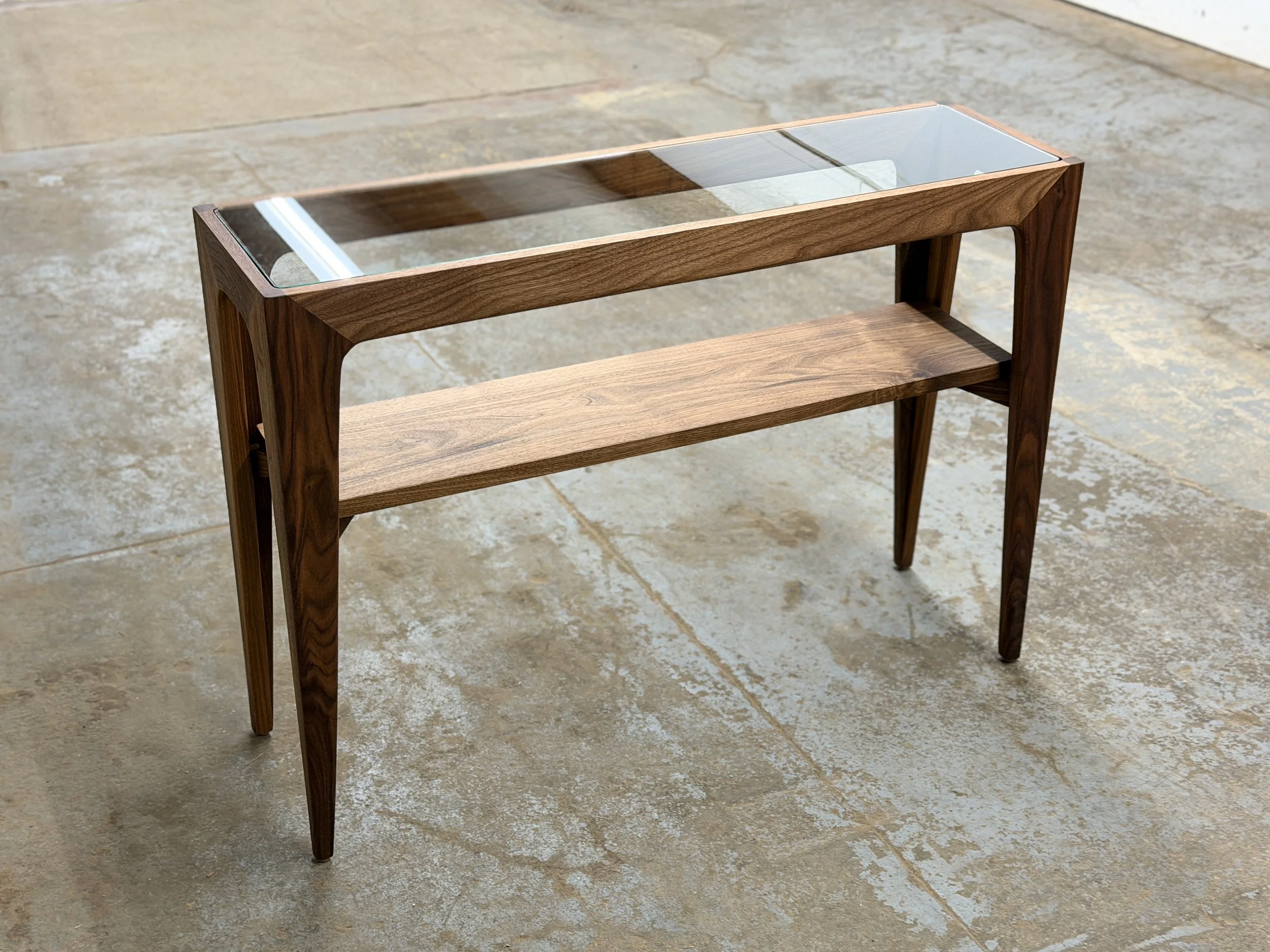 A wooden and glass console table with a lower wooden shelf on four tapered legs, set on a concrete floor.