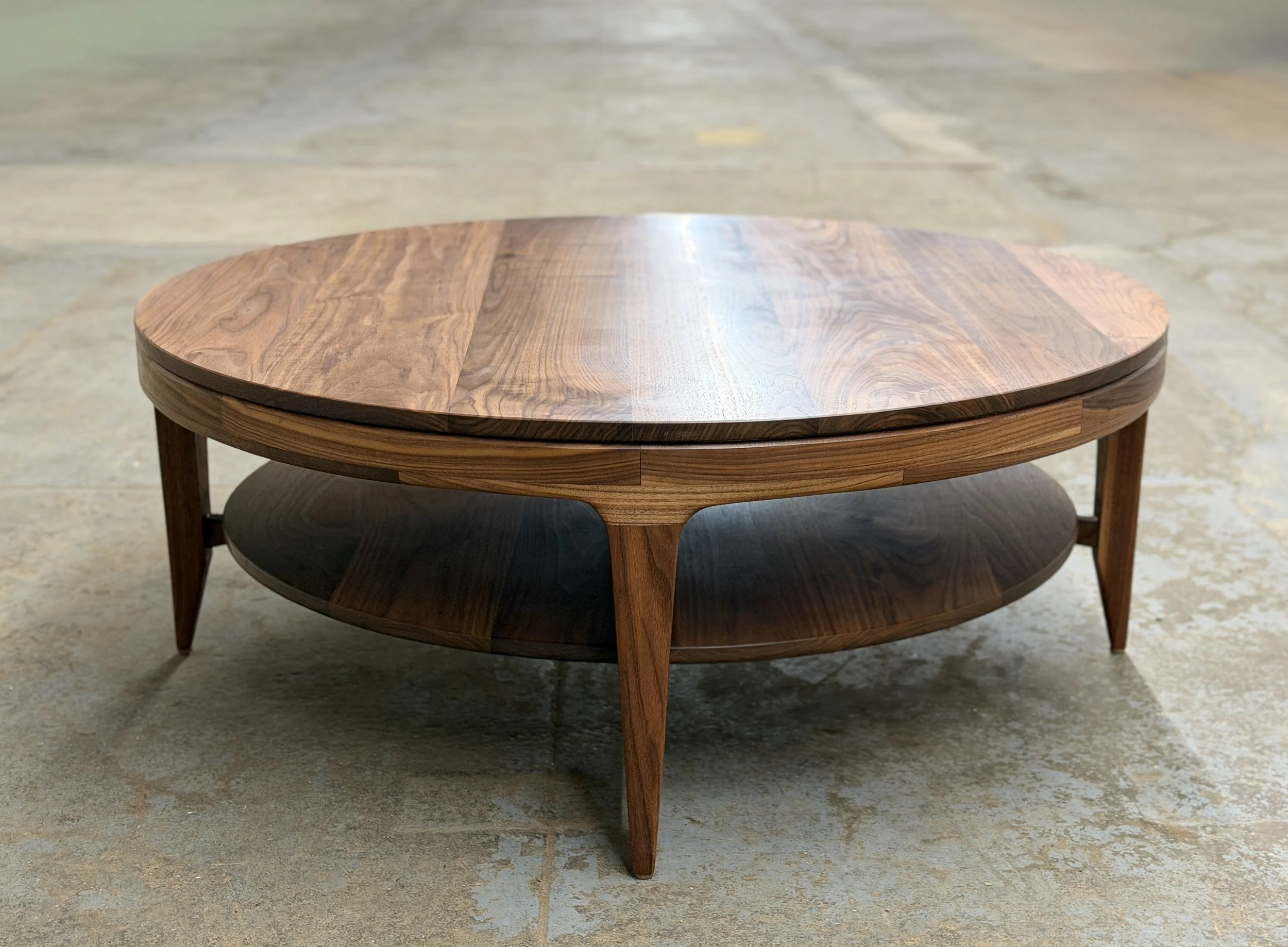 Wooden coffee table with a round top and lower shelf, placed on a concrete floor.