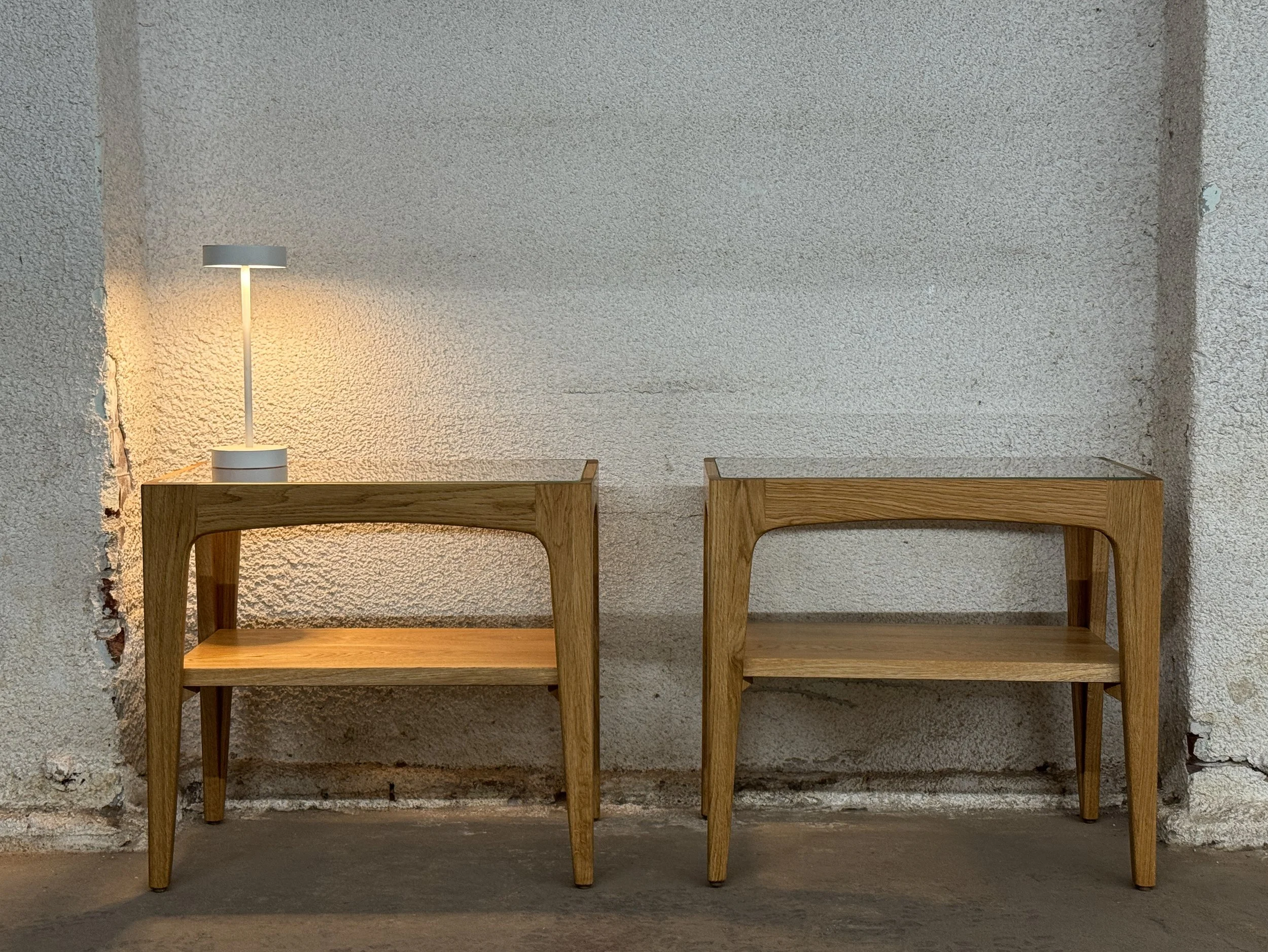 Two wooden side tables with curved edges and lower shelves, placed against a textured white wall. The left table has a white modern desk lamp turned on.