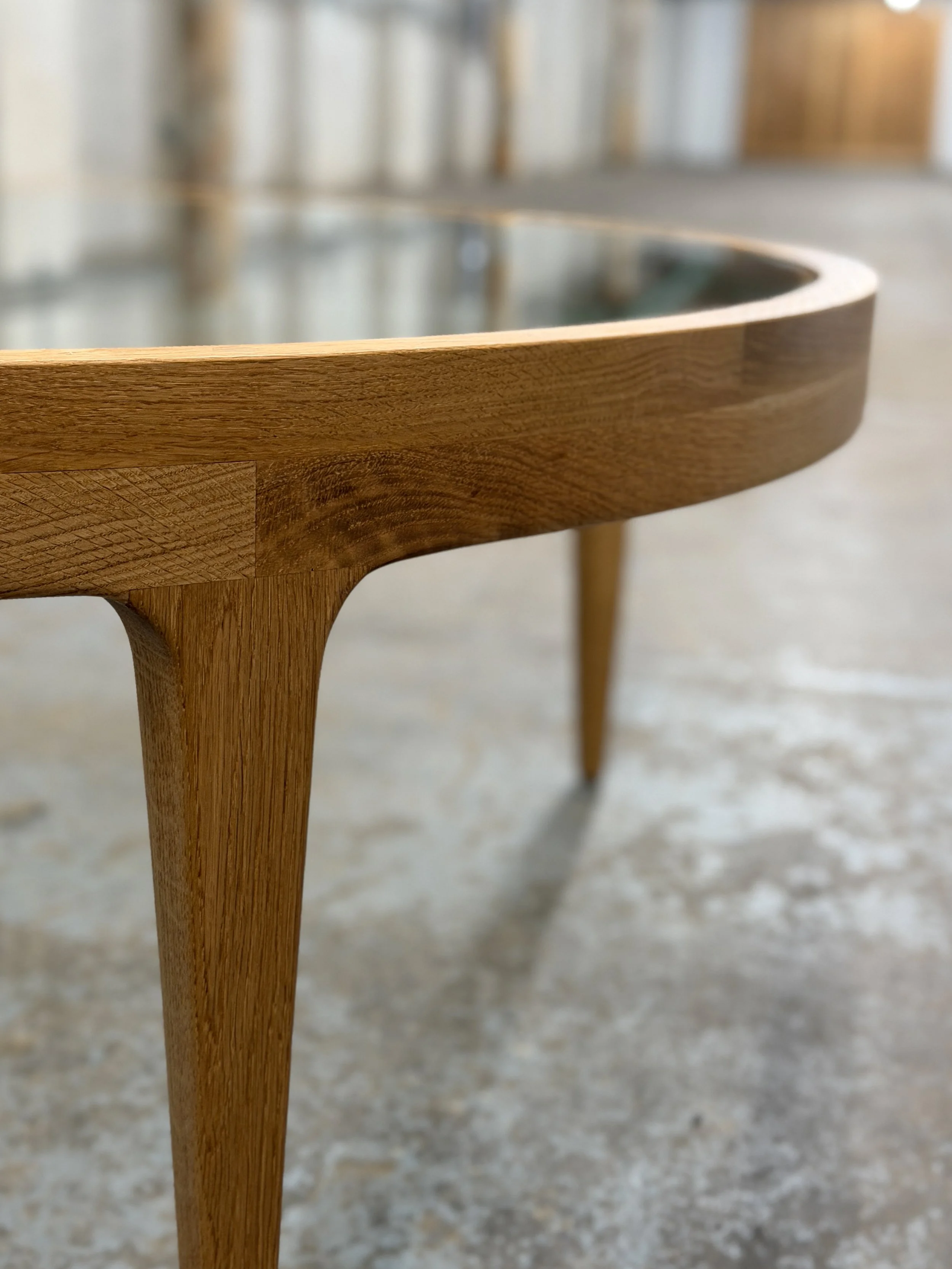 Close-up view of a wooden furniture piece, possibly a chair or table, with a curved edge and four tapered legs, in a well-lit indoor space with a concrete or stone floor.