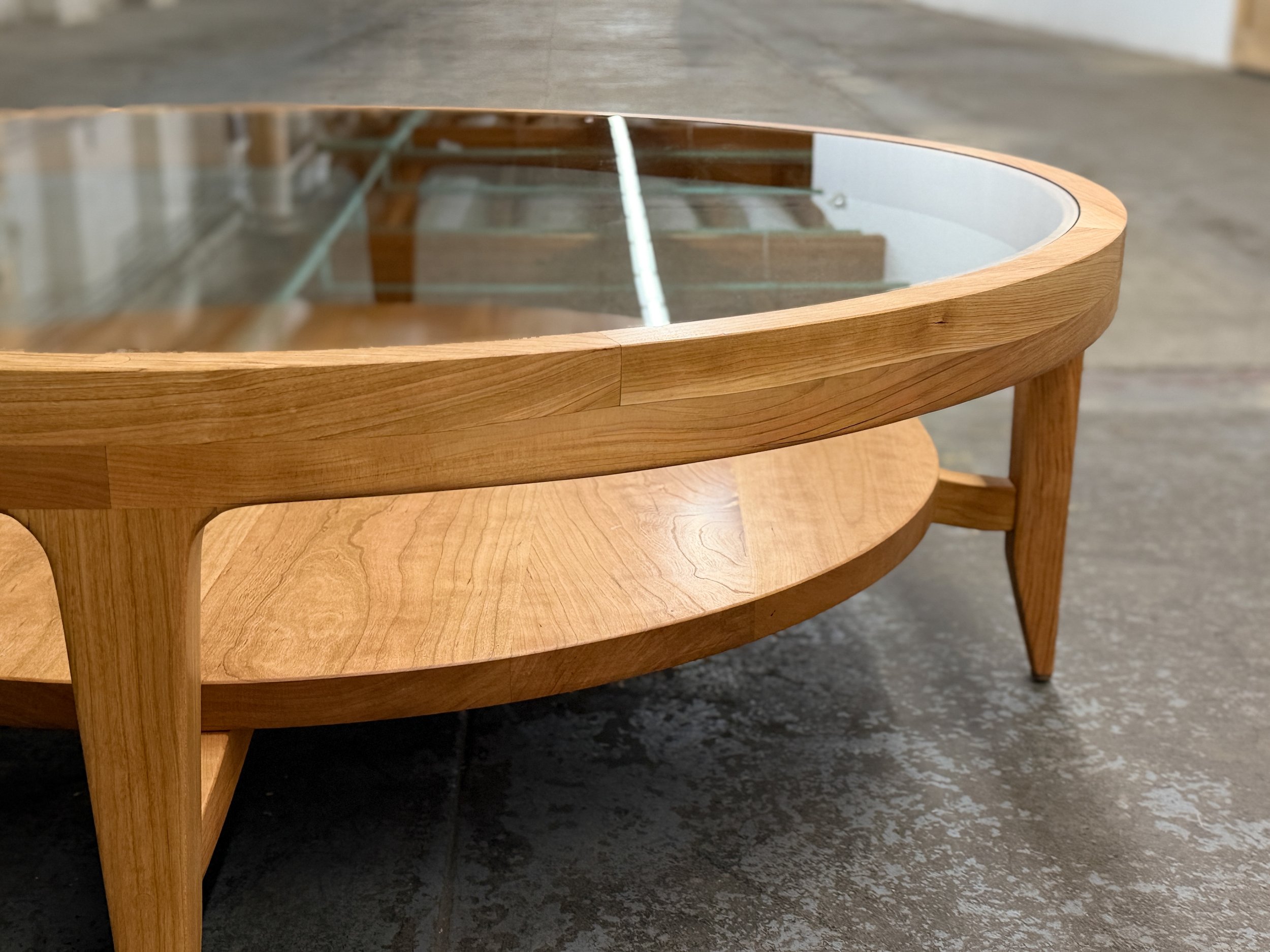 Wooden oval coffee table with glass top and lower shelf, placed on a concrete floor.