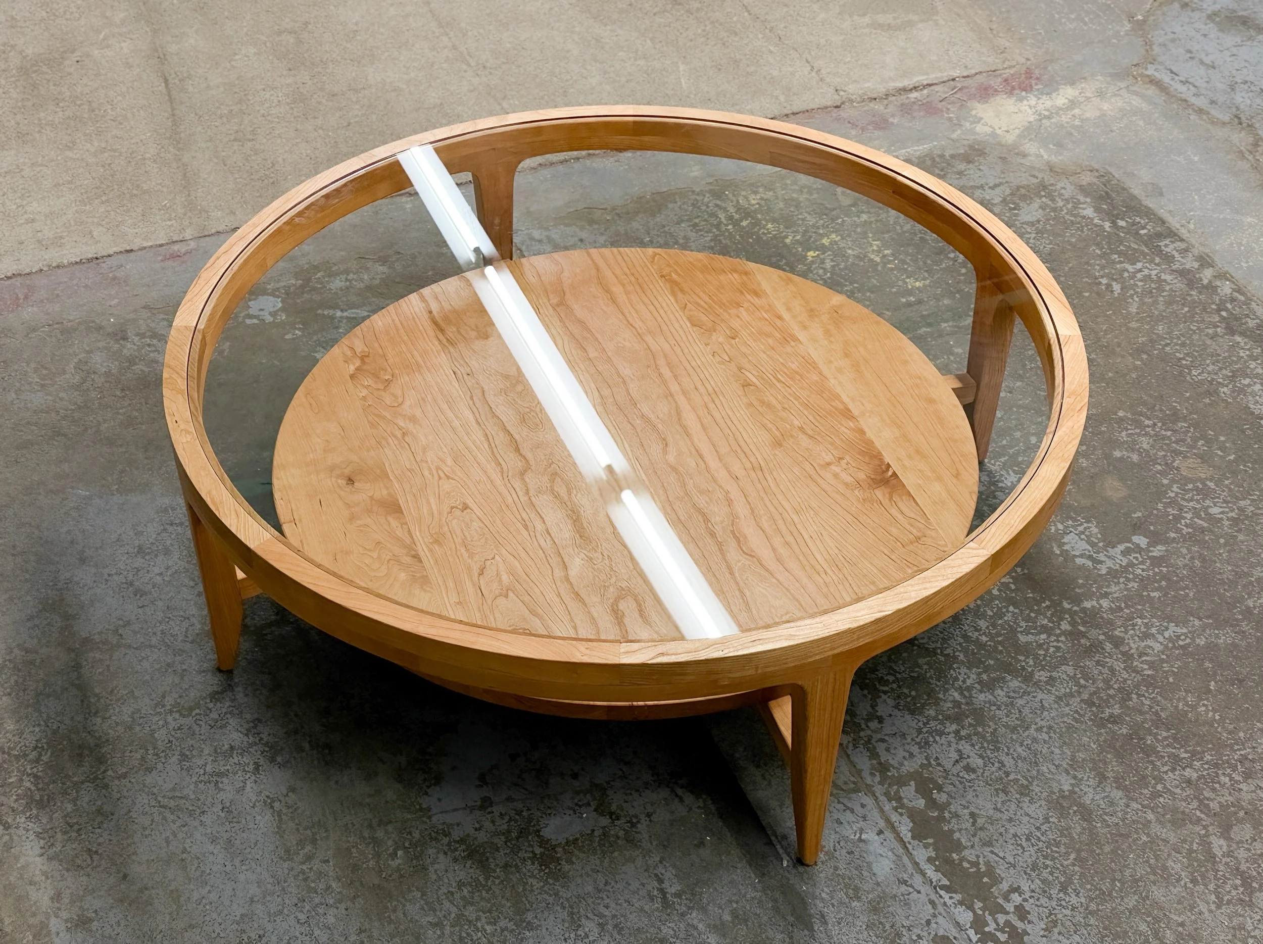 A round wooden coffee table with a glass top, placed on a concrete floor.