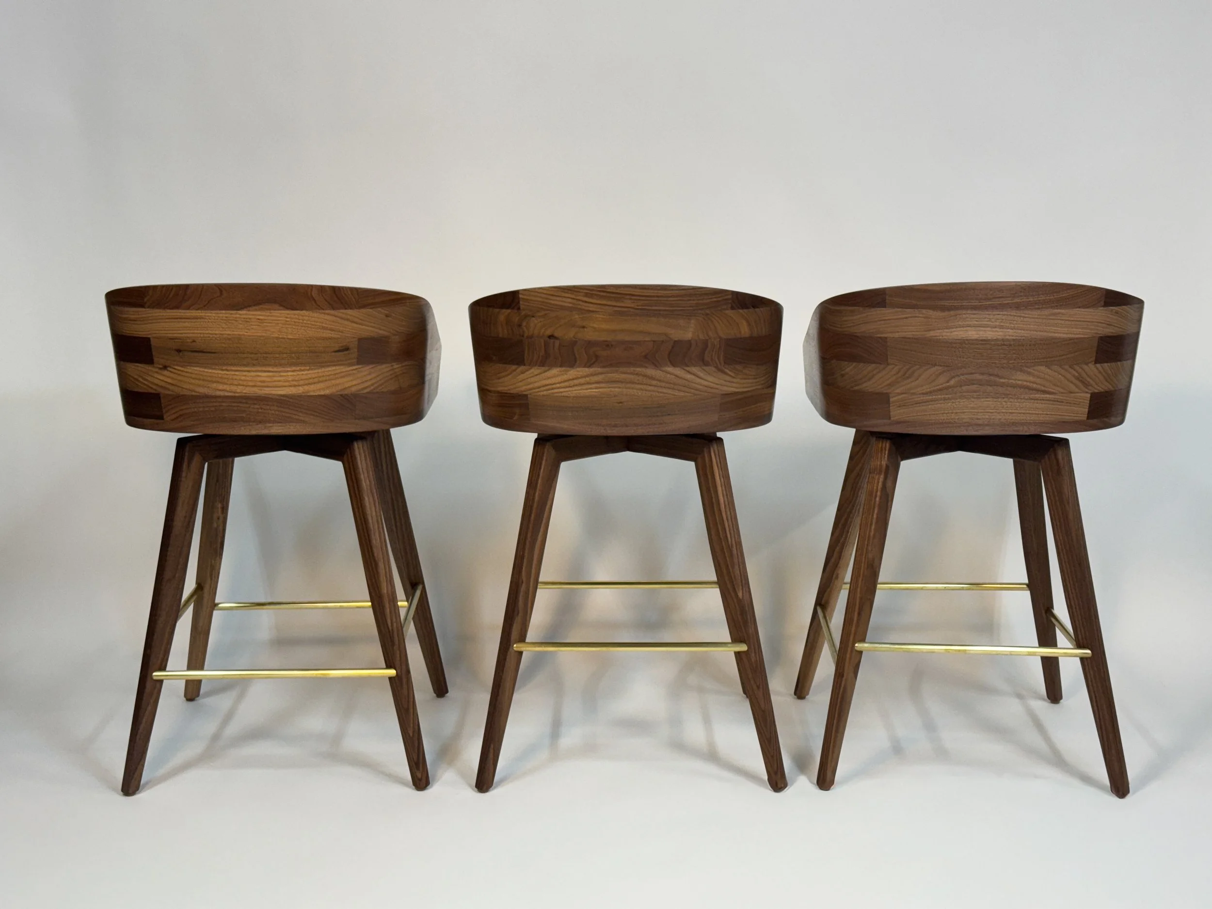 Three wooden bar stools with curved backs and four angled legs each, connected with horizontal gold-colored bars, set against a plain white background.