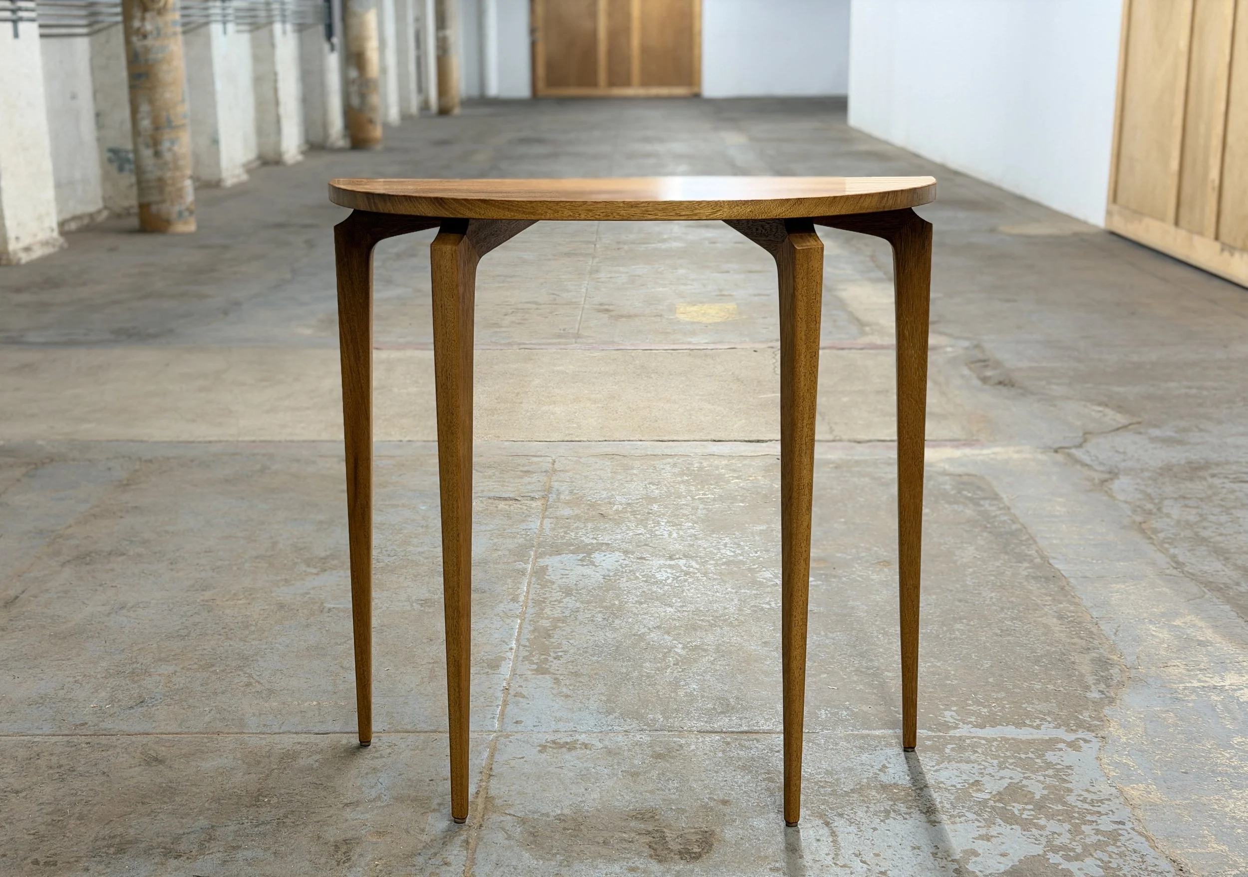 A wooden table with four slender legs is in the center of a rustic, unfinished indoor space with concrete walls and floors.