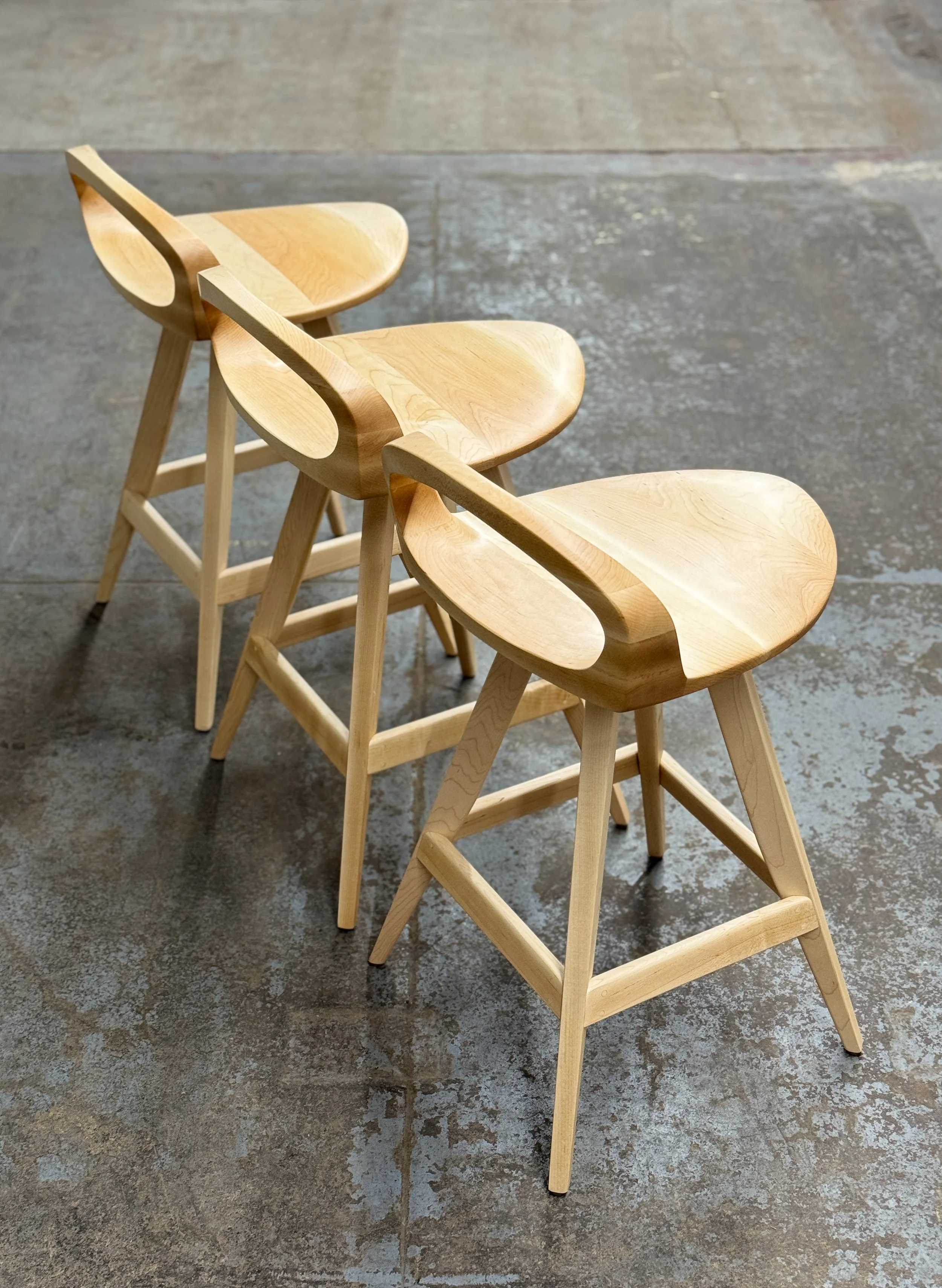 Three wooden chairs with curved seats and backrests, arranged in a line on a concrete floor.