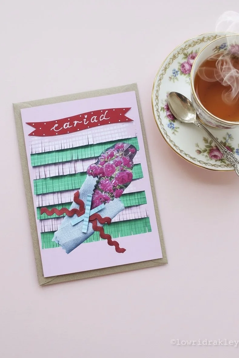 Handcrafted Welsh collage greeting card featuring a fuchsia rose bouquet and the word 'ariad,' pictured next to a streaming cup of tea in a vintage floral teacup.
