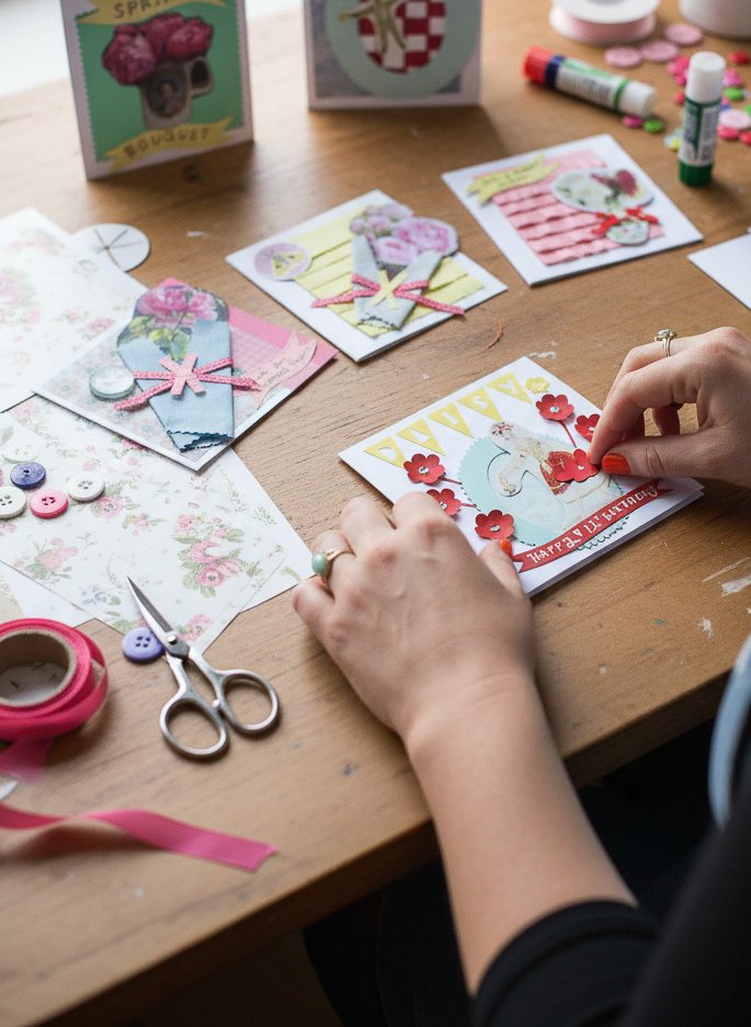 Artist Lowri Drakley's hands carefully placing a red paper flower onto a hand-cut collage card on a wooden studio table covered in vintage papers, ribbons, and scissors.