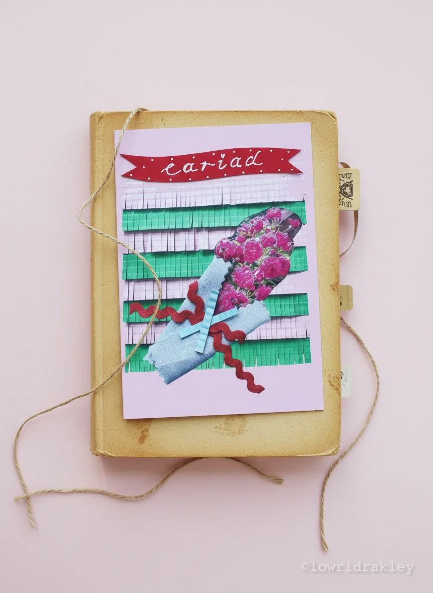 A handmade "Cariad" greeting card featuring a 3D fuchsia floarl bouquet collage. The bouquet is wrapped in light blue paper with red rickracktrim and set against a background of pink and green fringed paper. The card rests on an antique book.