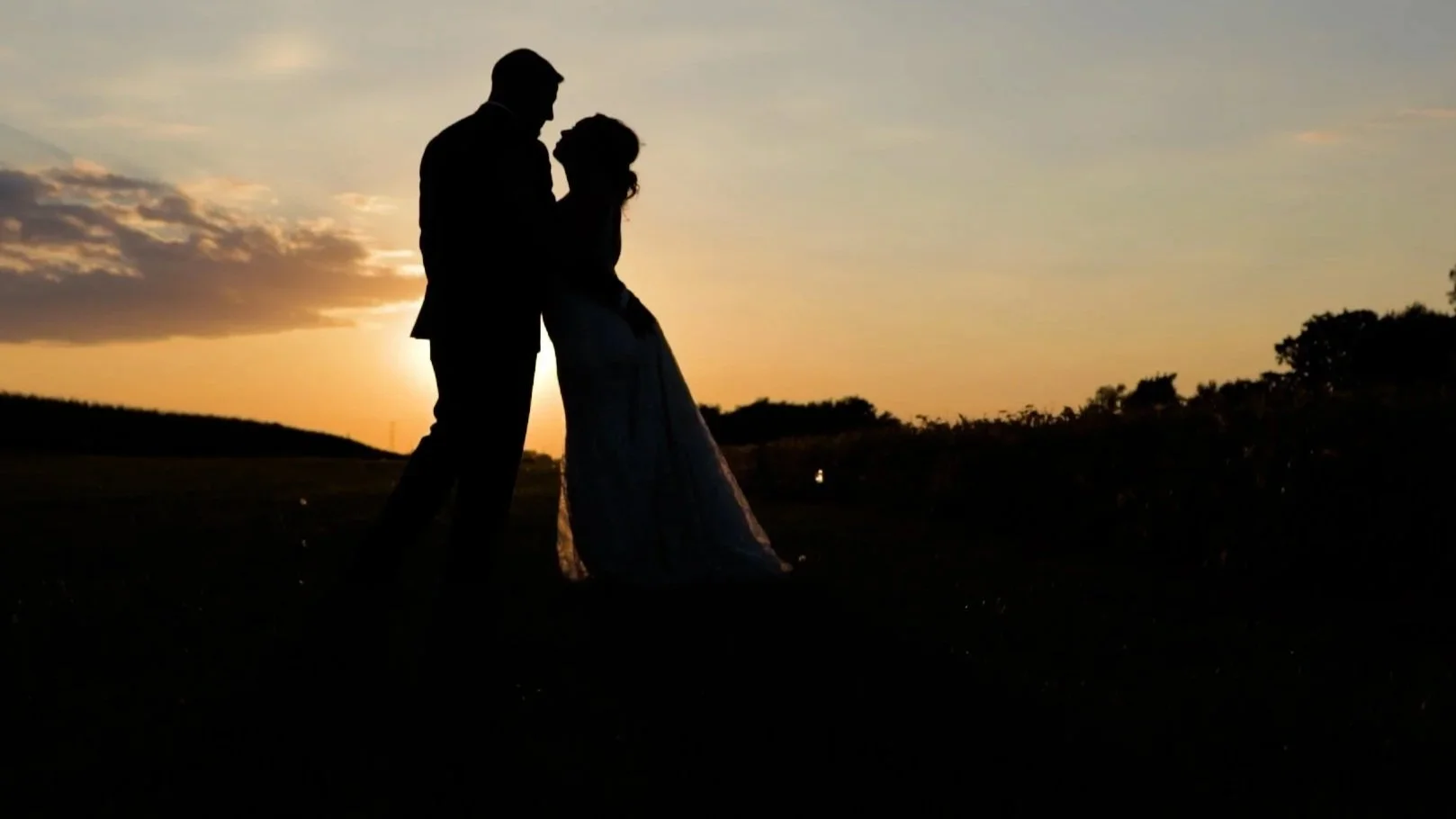 Silhouette of a couple dancing outdoors during sunset with a scenic landscape and cloudy sky in the background.