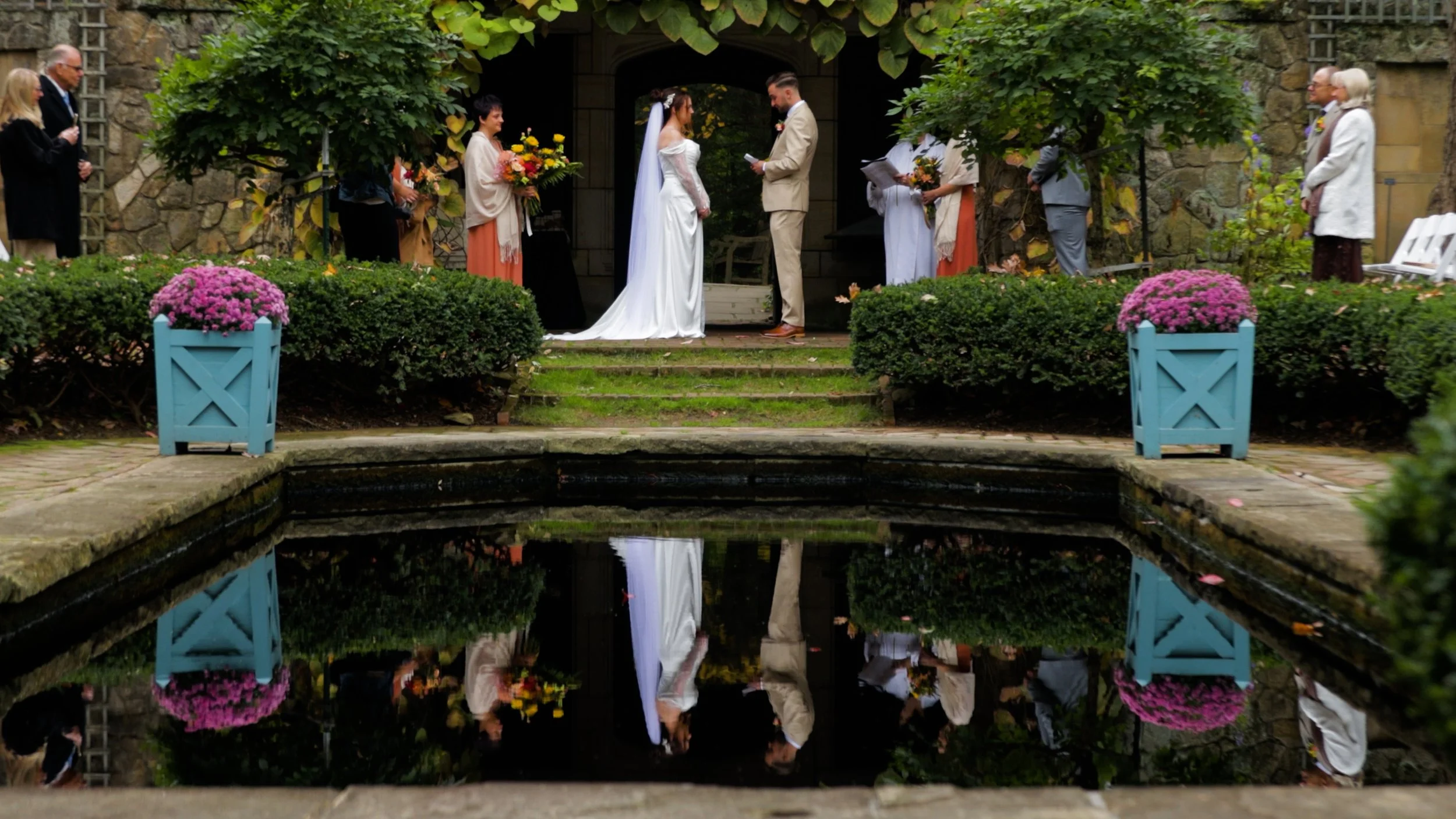 A wedding ceremony taking place at Stan Hywet Hall and Gardens at the Reflecting Pool. Outdoors in front of a stone building, with the bride and groom standing before officiants and guests.