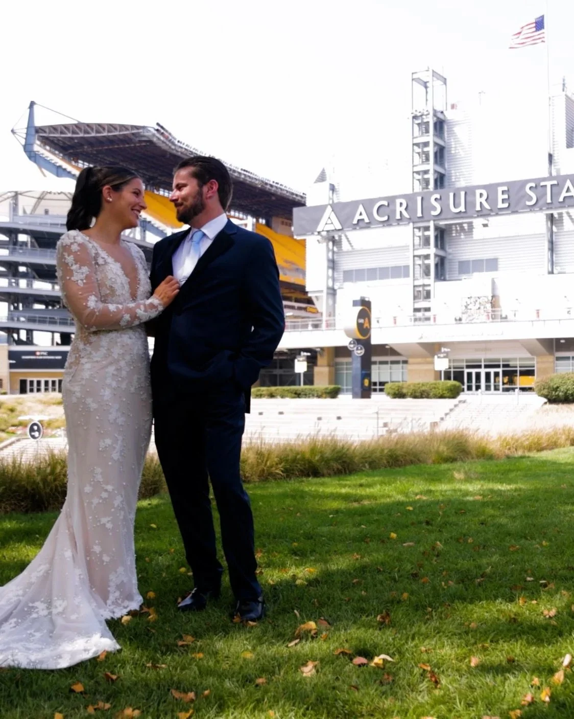 A couple in wedding attire standing on grass in front of a Acrisure Stadium in Pittsburgh Pa., smiling and looking at each other.