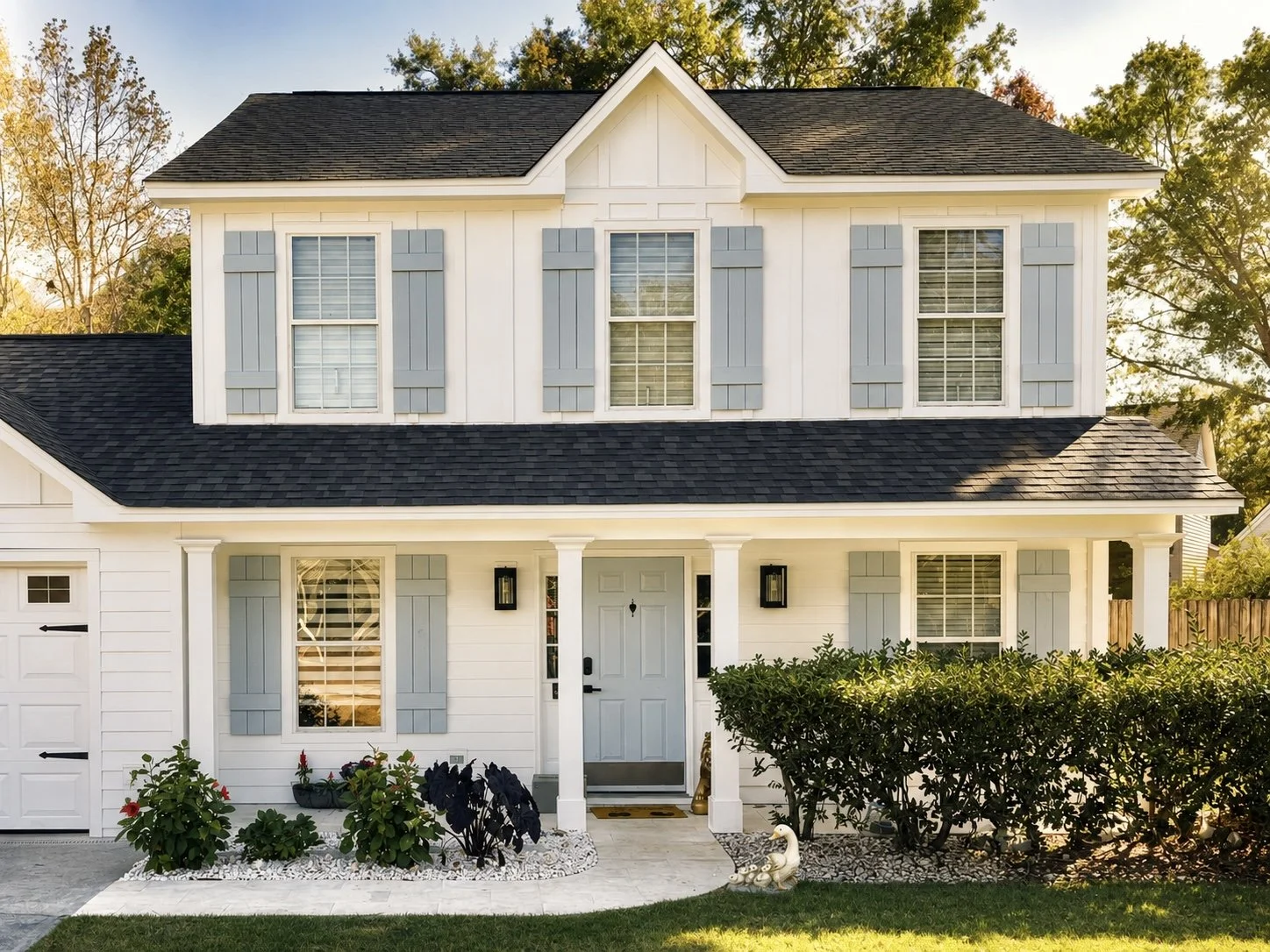 A white two-story house with light blue shutters, black roof, front porch with columns, and a small garden with shrubs and flowers.