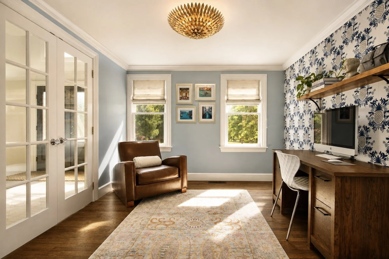 Bright living room with light blue walls, three windows with beige blinds, a leather armchair, a decorative rug, a wooden desk with a computer and a white chair, a wall-mounted shelf with decorative items, and a floral wallpaper accent wall.