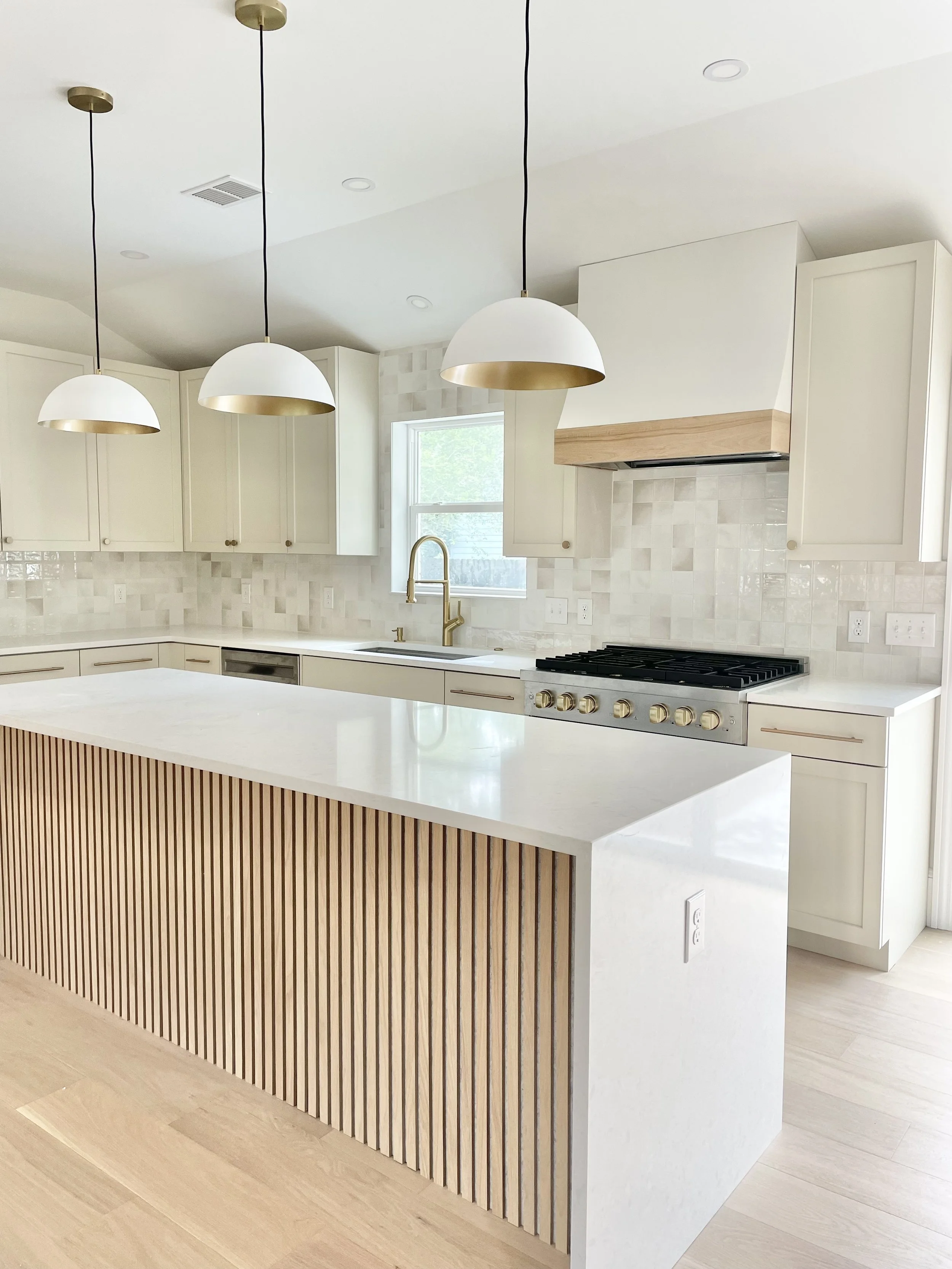 Modern kitchen with white cabinets, a white island with a wood slatted front, pendant lights, a window, and a stove with a wooden range hood.