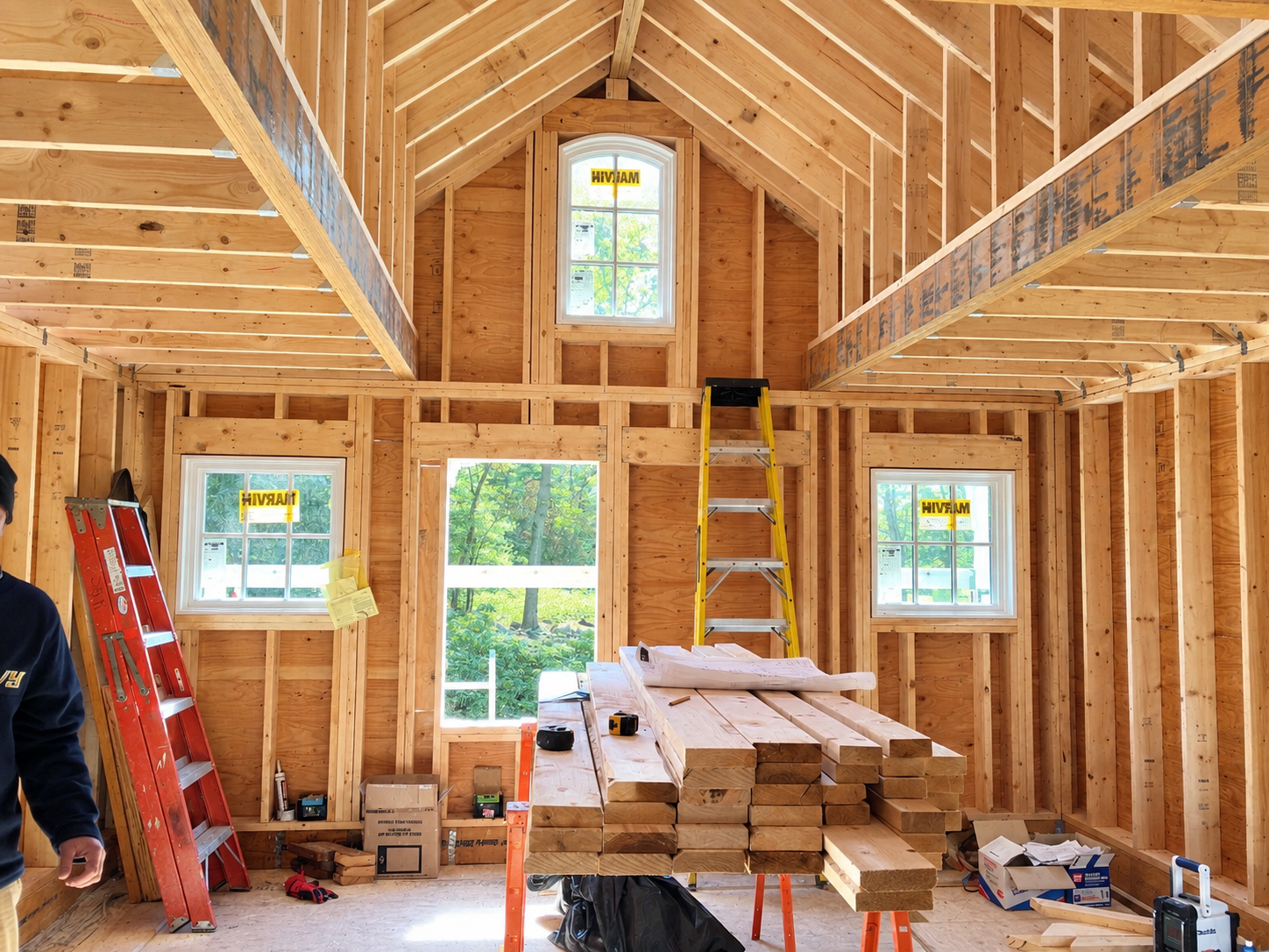 Interior of a house under construction with wood framing, windows, and construction tools, including a ladder, a stack of lumber, and a work table.