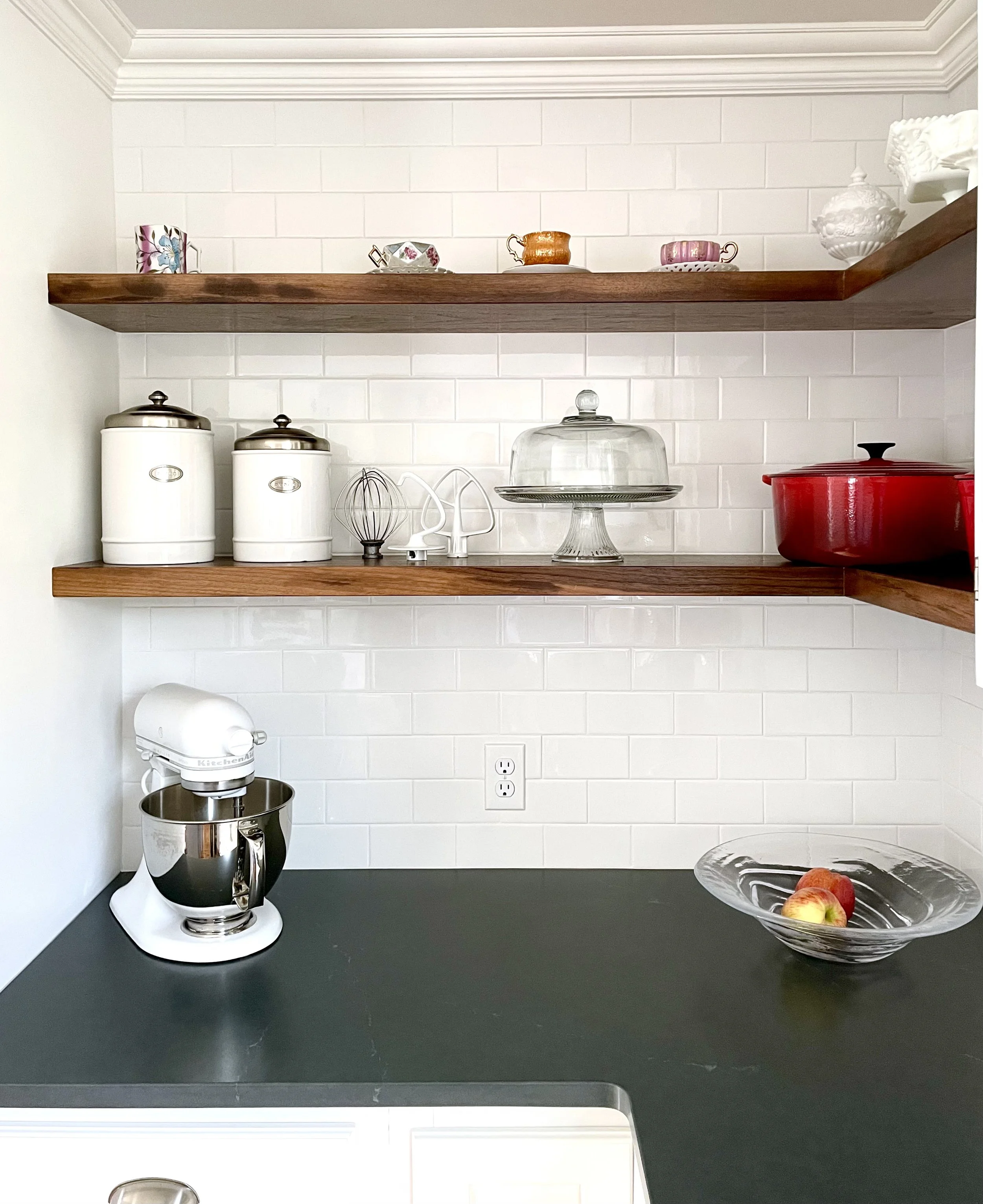 Kitchen countertop with a slate surface, a stand mixer with a metal mixing bowl, a glass cake stand empty, a glass bowl with apples, and a glass dish with additional apples. Two wooden shelves above hold decorative teacups, saucers, and a white vase.