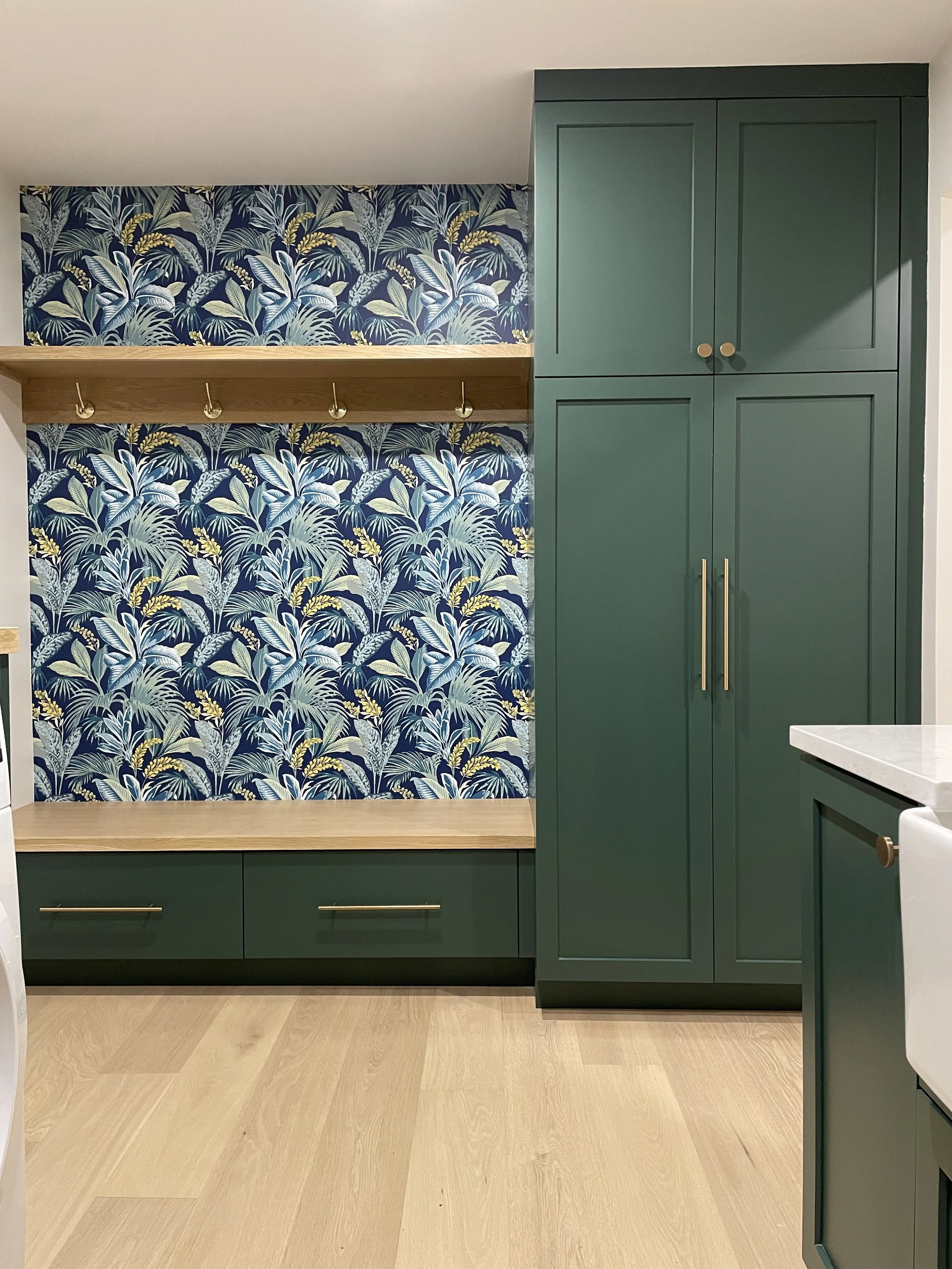 A modern kitchen corner with green cabinets, a wooden shelf with hooks, a patterned wallpaper with leaf designs, and a light hardwood floor.