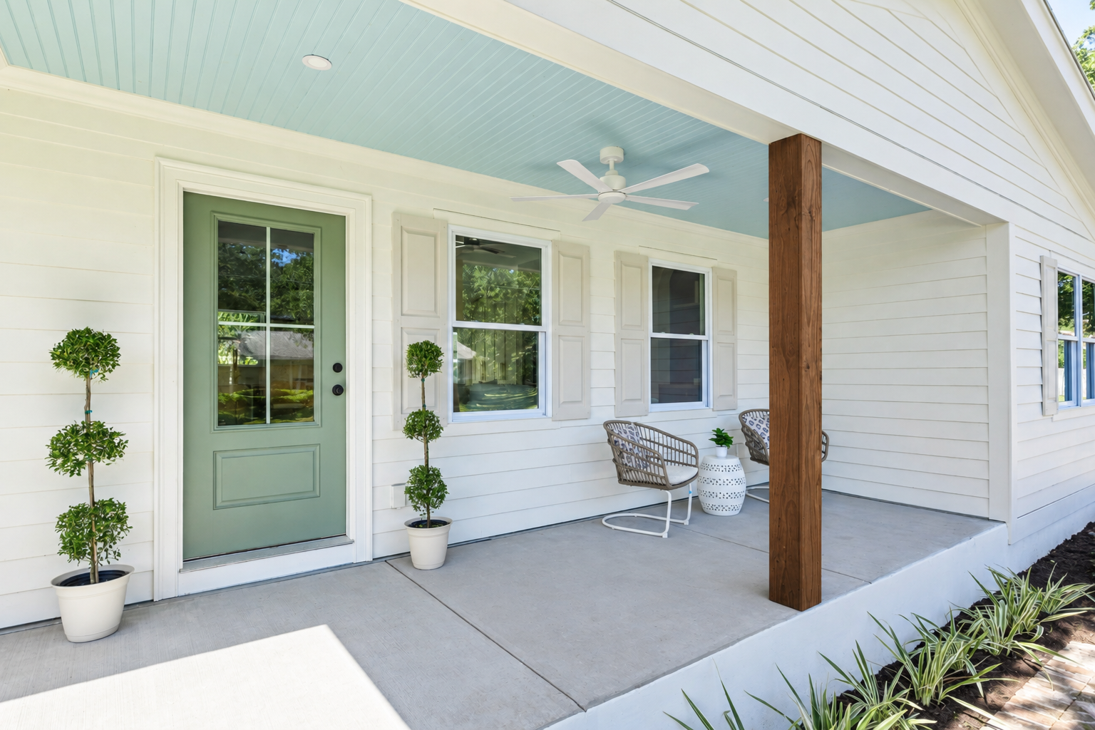 Front porch of a house with a green door, two potted topiary plants, two chairs, a small white side table, and a ceiling fan, with a concrete floor and siding.