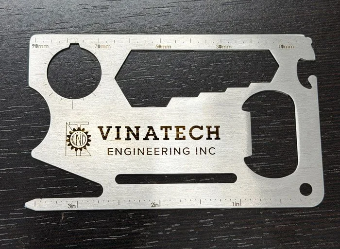 Metal multi-tool with cutouts and measurements, engraved with 'VINATECH ENGINEERING INC' and a gear logo, on a black surface.
