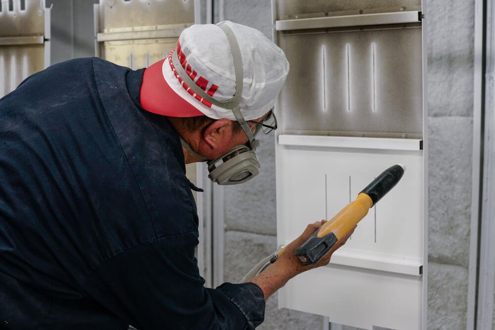 A worker wearing a cap, mask, and glasses using a spray gun or paint sprayer on a metal surface in an industrial setting.