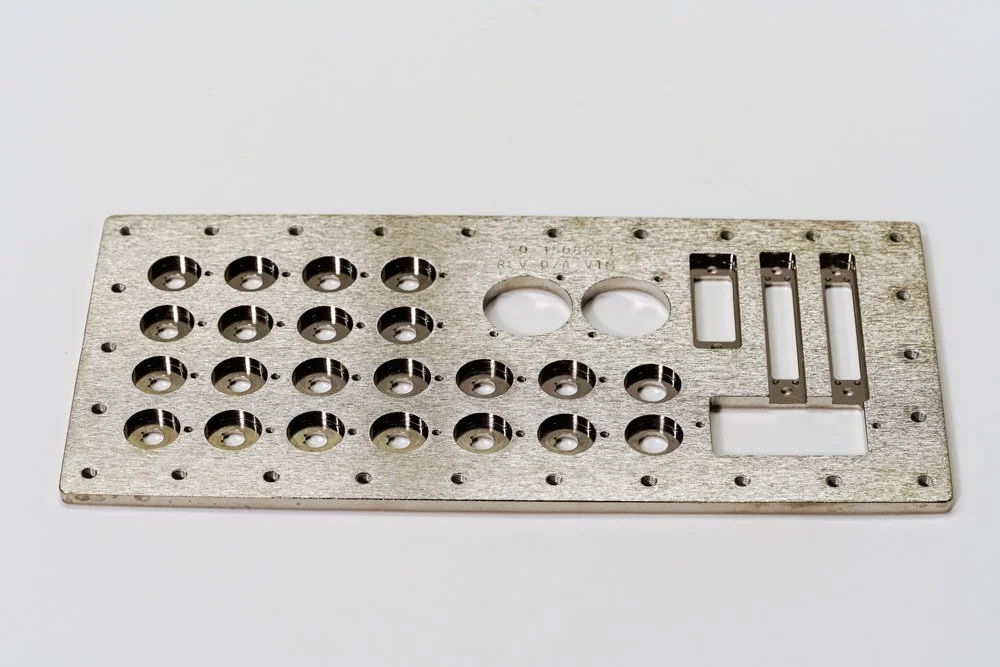 A rectangular metal plate with multiple circular holes, a few larger holes, and some rectangular cutouts, placed on a white surface.