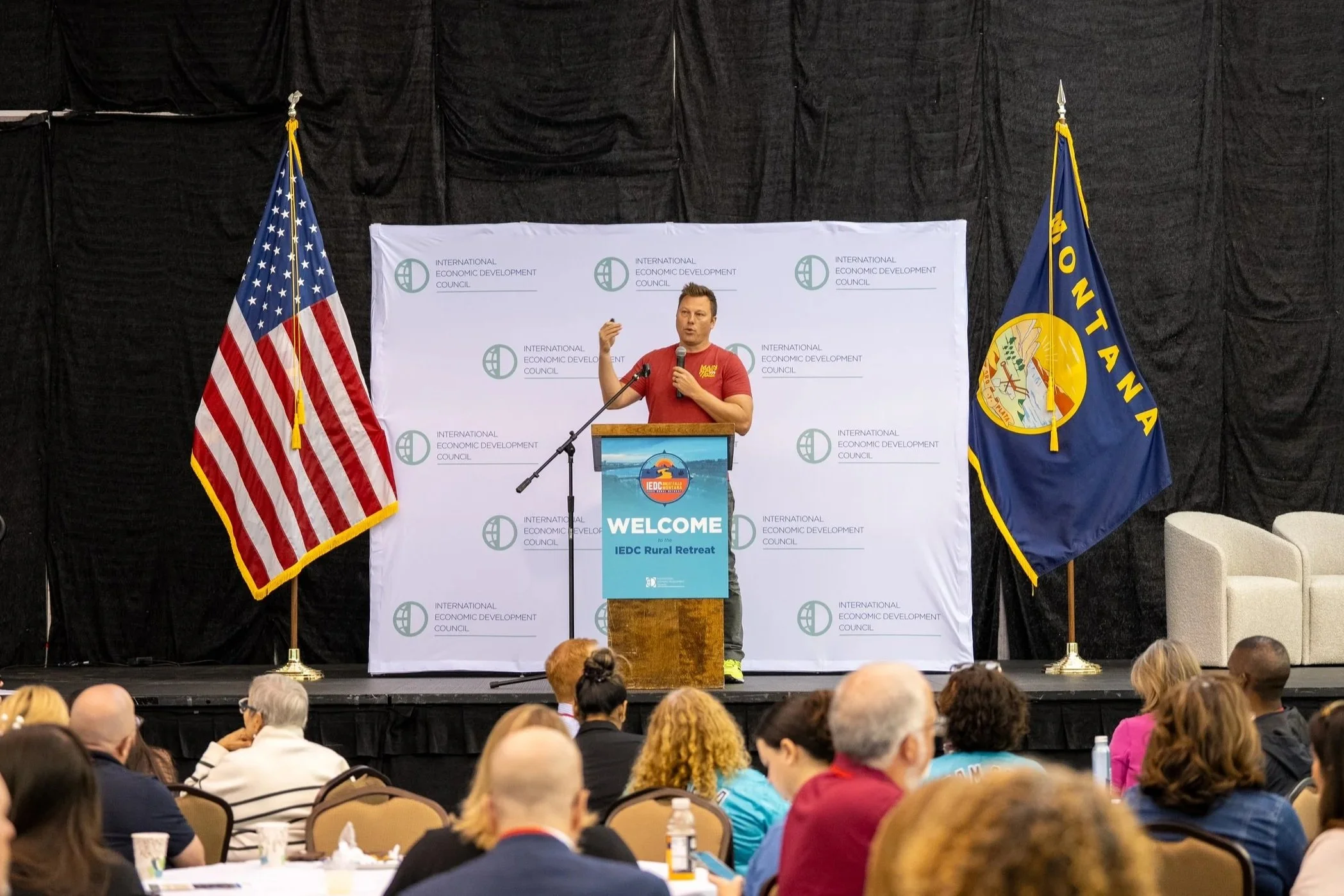 A man speaking at a podium during a conference, with American and Montana flags on either side, and a backdrop with the International Economic Development Council logo. The audience is seated in front.