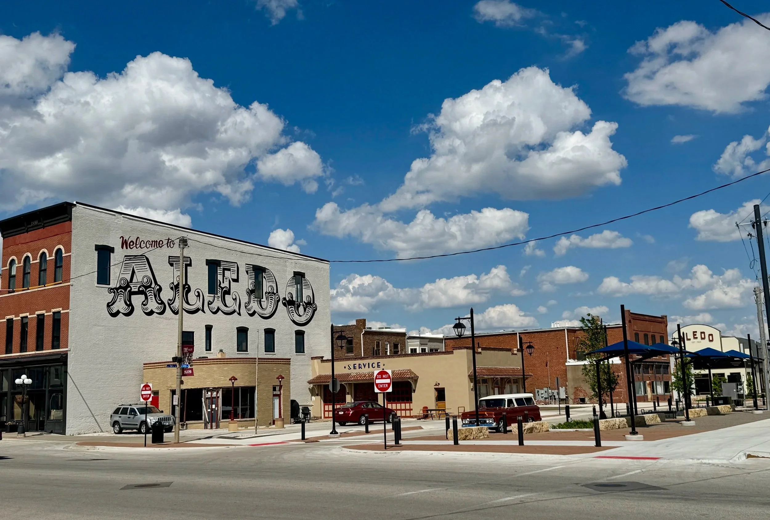 A small town square with brick buildings, including a large white building with a mural that says 'Welcome to ALLED', under a blue sky with scattered clouds.