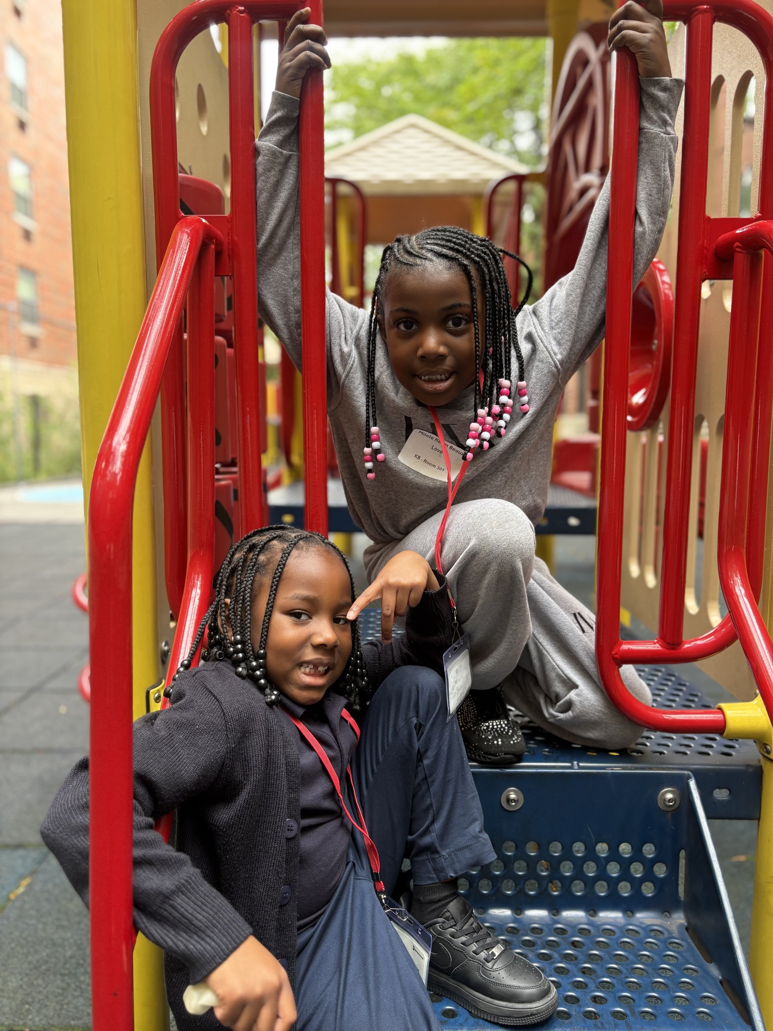 Two young girls with braided hair and dark clothing playing on a colorful playground structure outdoors.