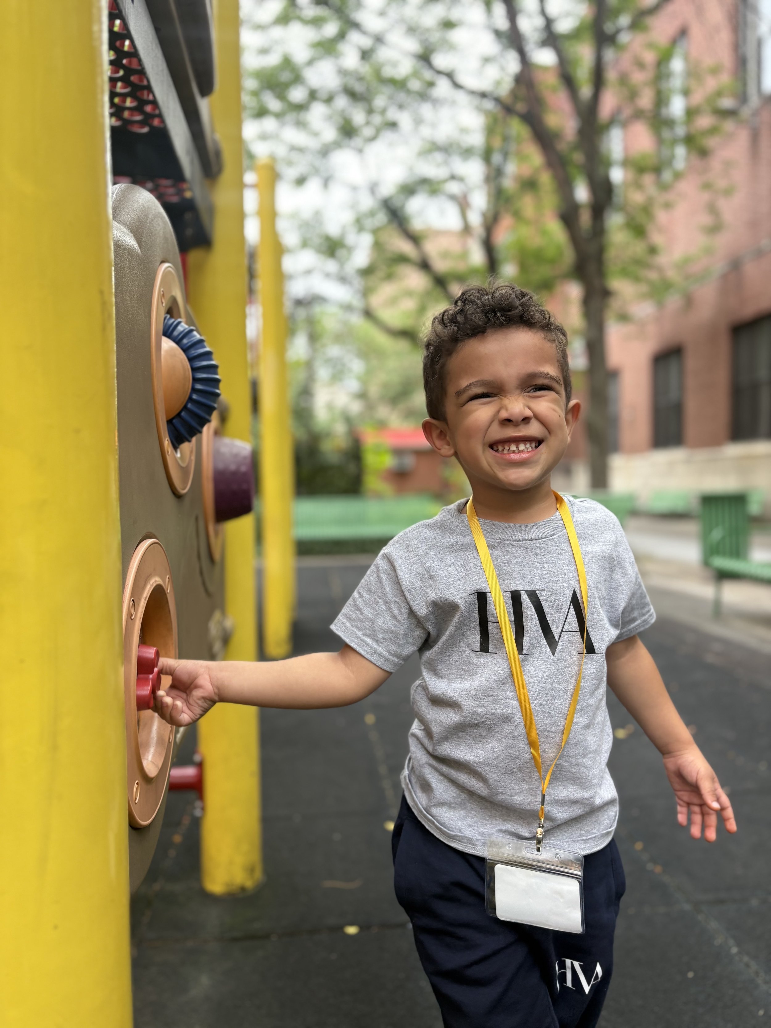 A young boy smiling at the camera while playing on an outdoor playground with colorful climbing panels, wearing a gray t-shirt, navy shorts, and a yellow lanyard with an ID badge around his neck.