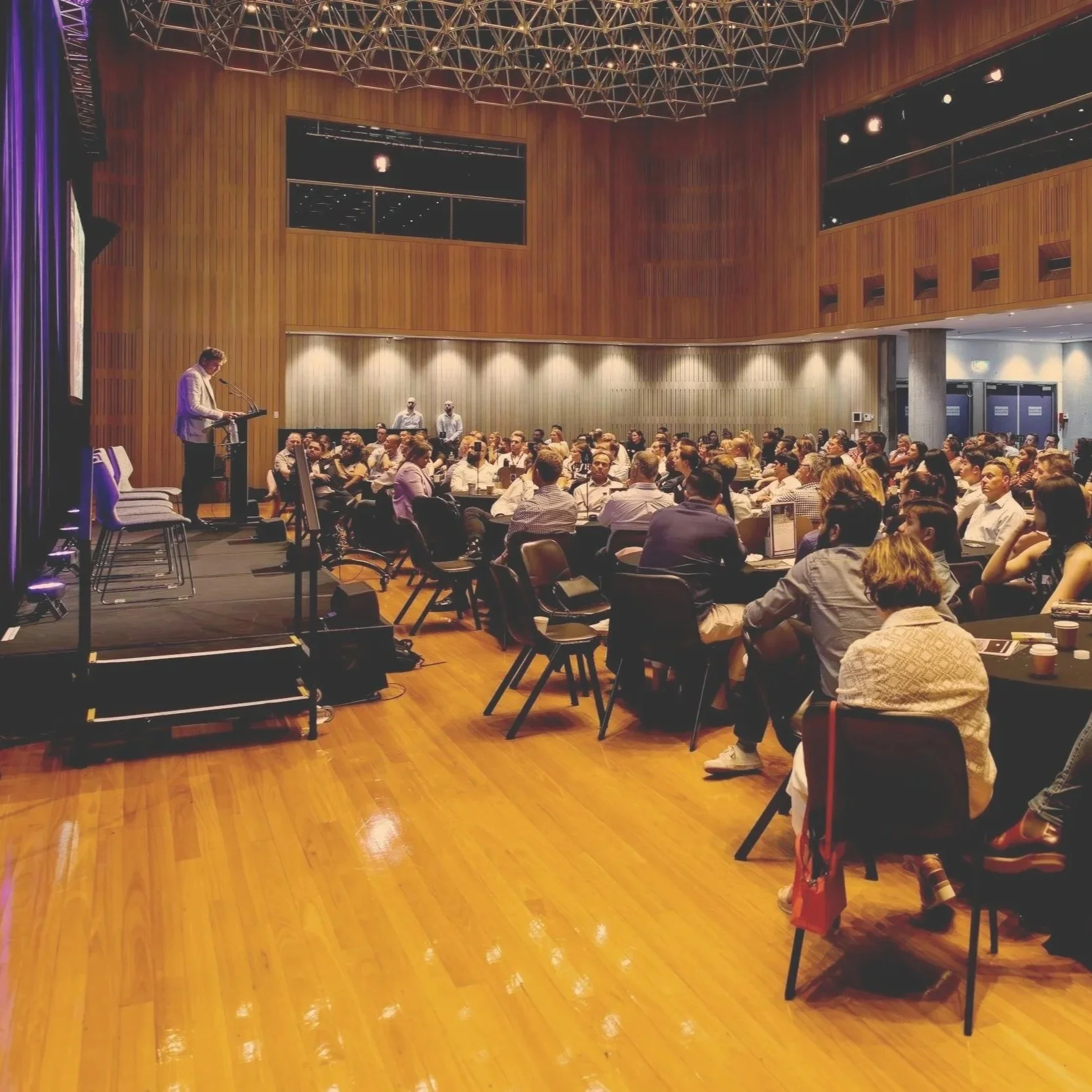 A man giving a presentation on stage to a large audience seated at round tables in a conference room with wooden walls and high ceiling.