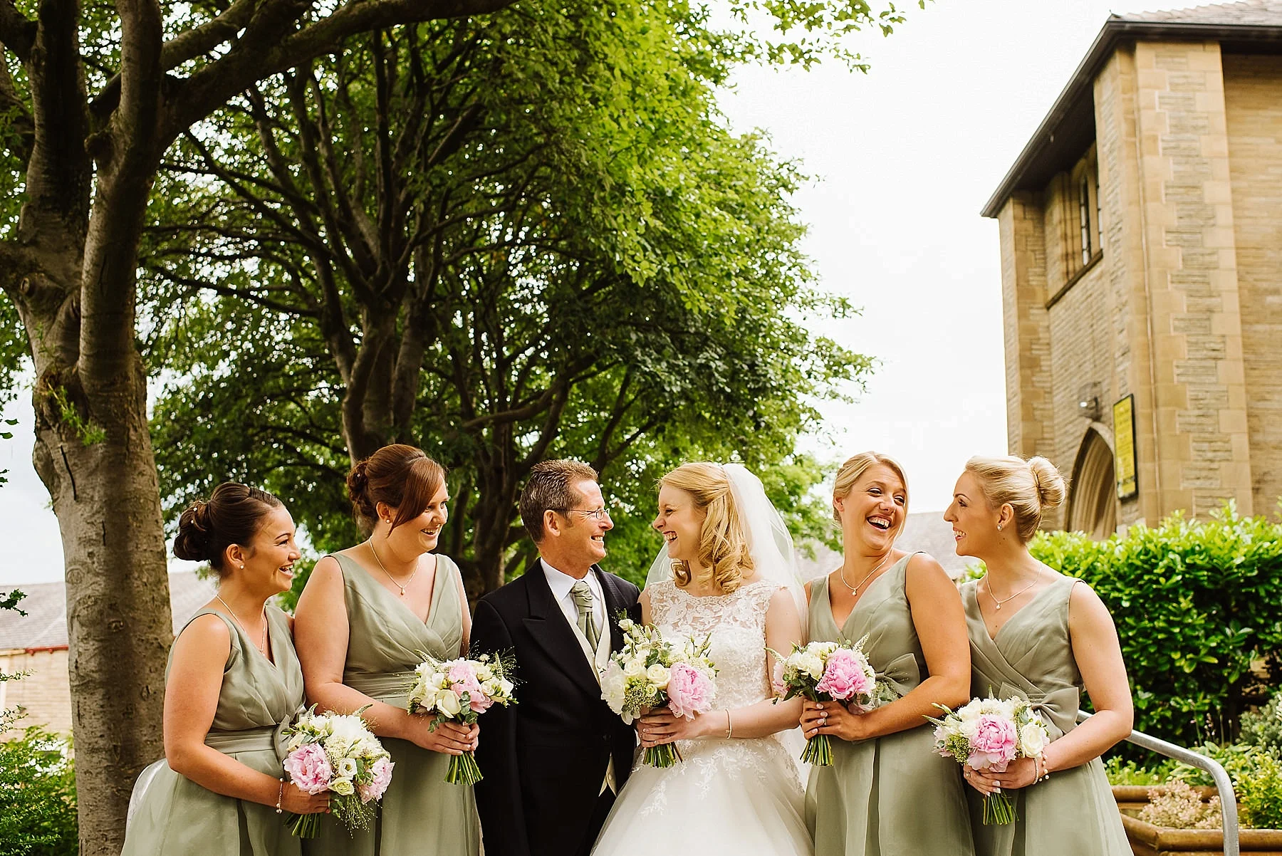 a bride with her father and four bridesmaids laughing outside a St Mary Magdalens C-of-E Church