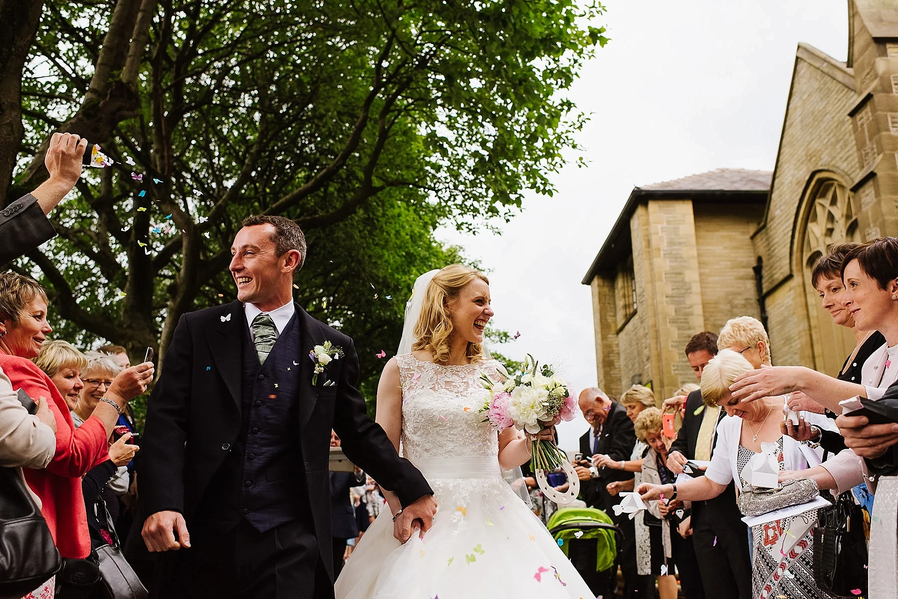 The bride and groom leaving the church grounds whilst they are lined up on either side by all their guests who are throwing confetti on them