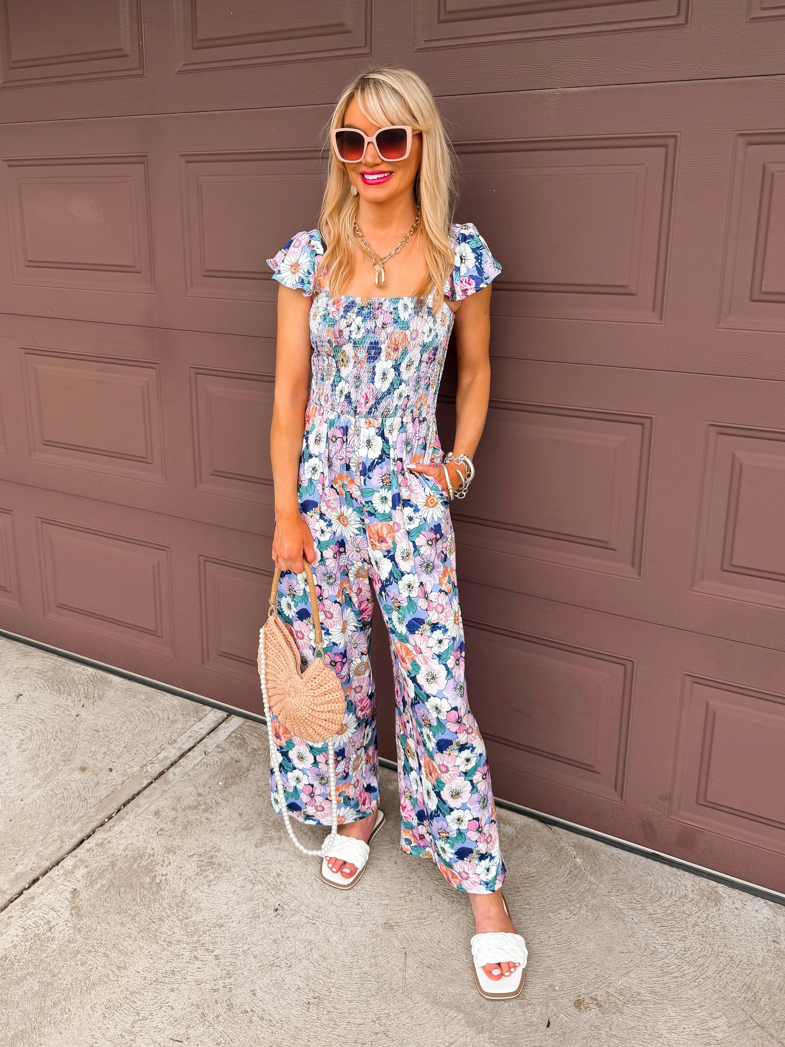blonde woman wearing stretchy smocked floral jumpsuit with flutter sleeves by Ekouear on Amazon with a seashell handbag and white braided sandal slides
