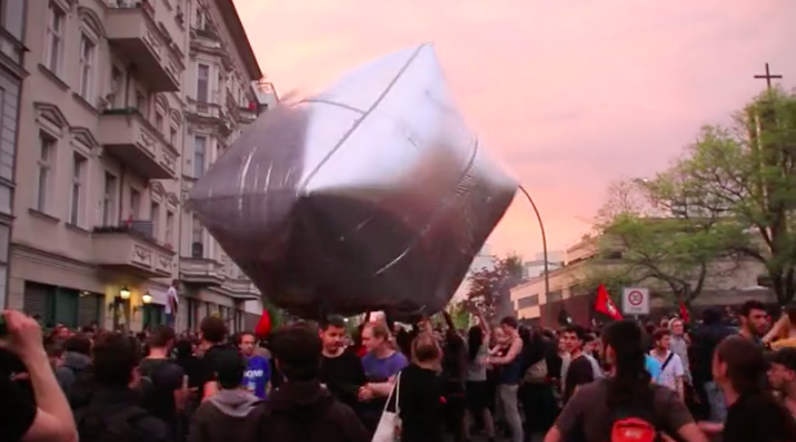 Screen grab from Inflatable Cobblestones Berlin Part 2 ( Vimeo ), by Artur (presumably Artúr van Balen), 2012 The video is captioned: On the 25th revolutionary 1st of May demonstration in Berlin-Kreuzberg, protesters were throwing huge inflatable cobblestones, made of silver-reflective foil and tape. The creative intervention was initiated by the art-activist collective “Eclectic Electric Collective” (EEC) and was meant as a celebration of an object which is both a symbol and a material weapon of anti-authoritarian struggle everywhere. It also aimed to bring new strategies of tactical frivolity into the demonstration. http://eclectic-electric-collective.blogspot.de/2012/05/under-pavement-beach-gigantic.html