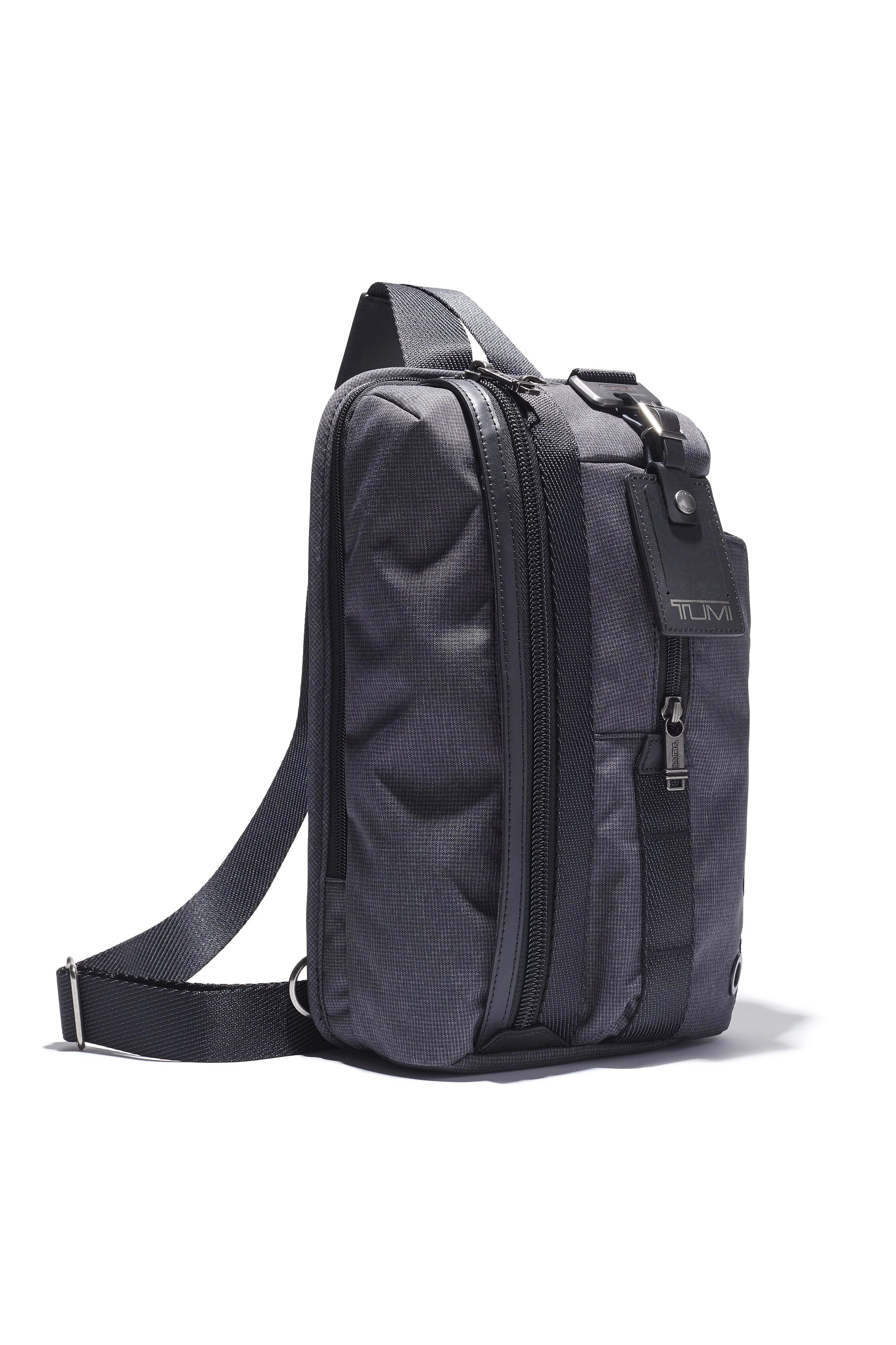 A dark gray backpack with black zippers and a black luggage tag hanging from the front, featuring the TUMI logo.