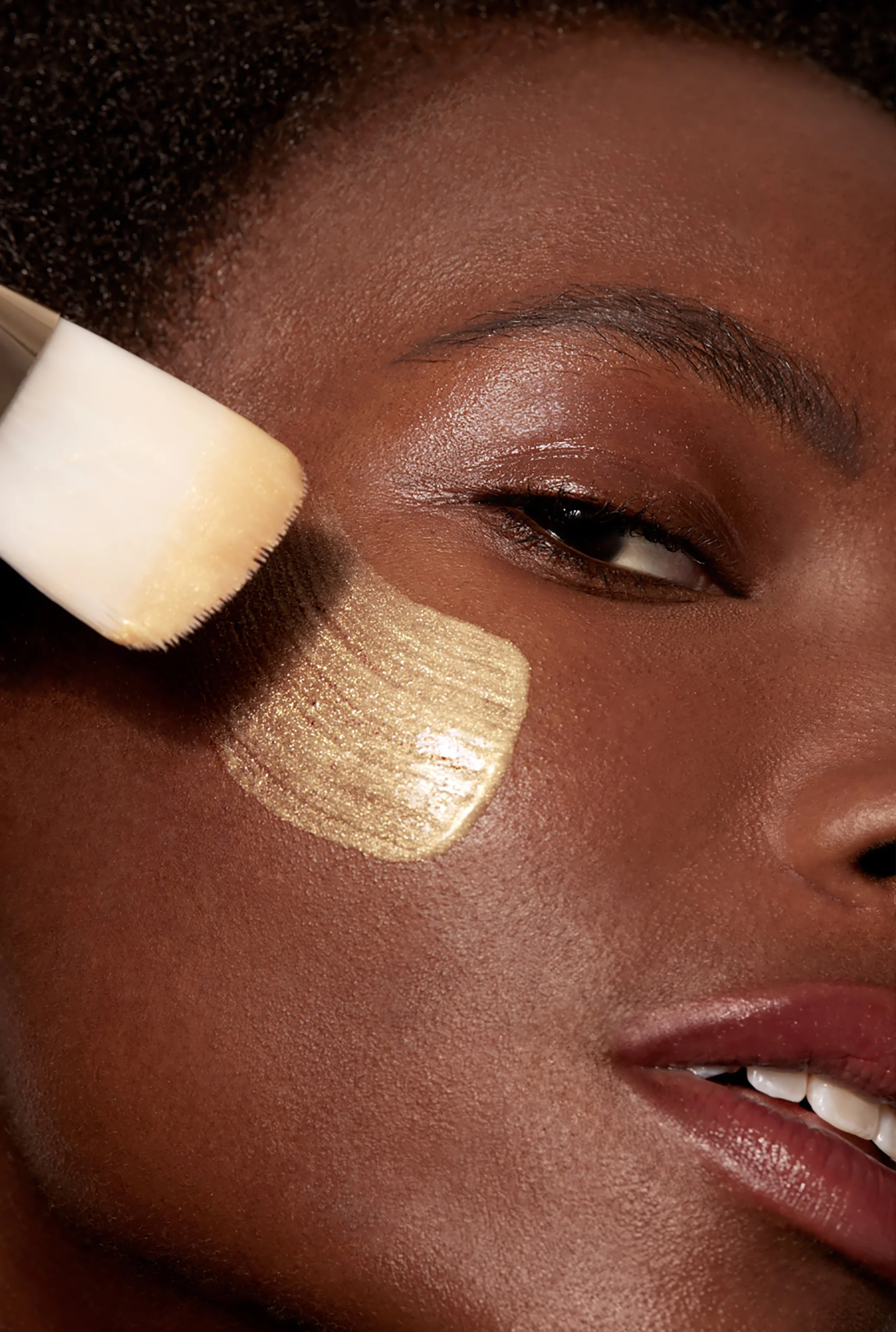A close-up of a woman with dark skin applying makeup using a white makeup sponge on her cheek. She has smooth skin, dark eyebrows, and a light lip color, with some shiny makeup visible on her cheek.
