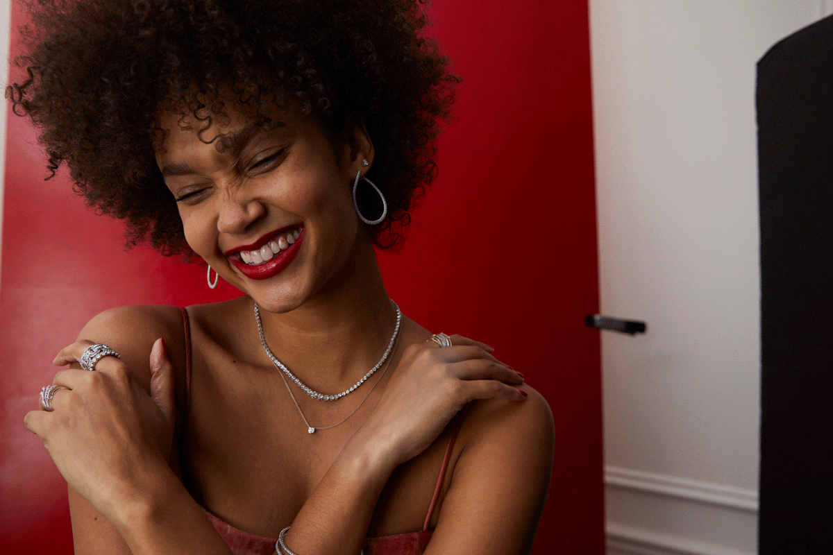 A woman with curly hair smiling and wearing jewelry against red and white background.