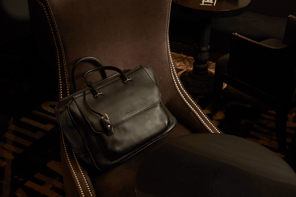 A black leather bag resting on a dark upholstered chair with diamond-tack detailing, in a dimly lit room with dark furniture.