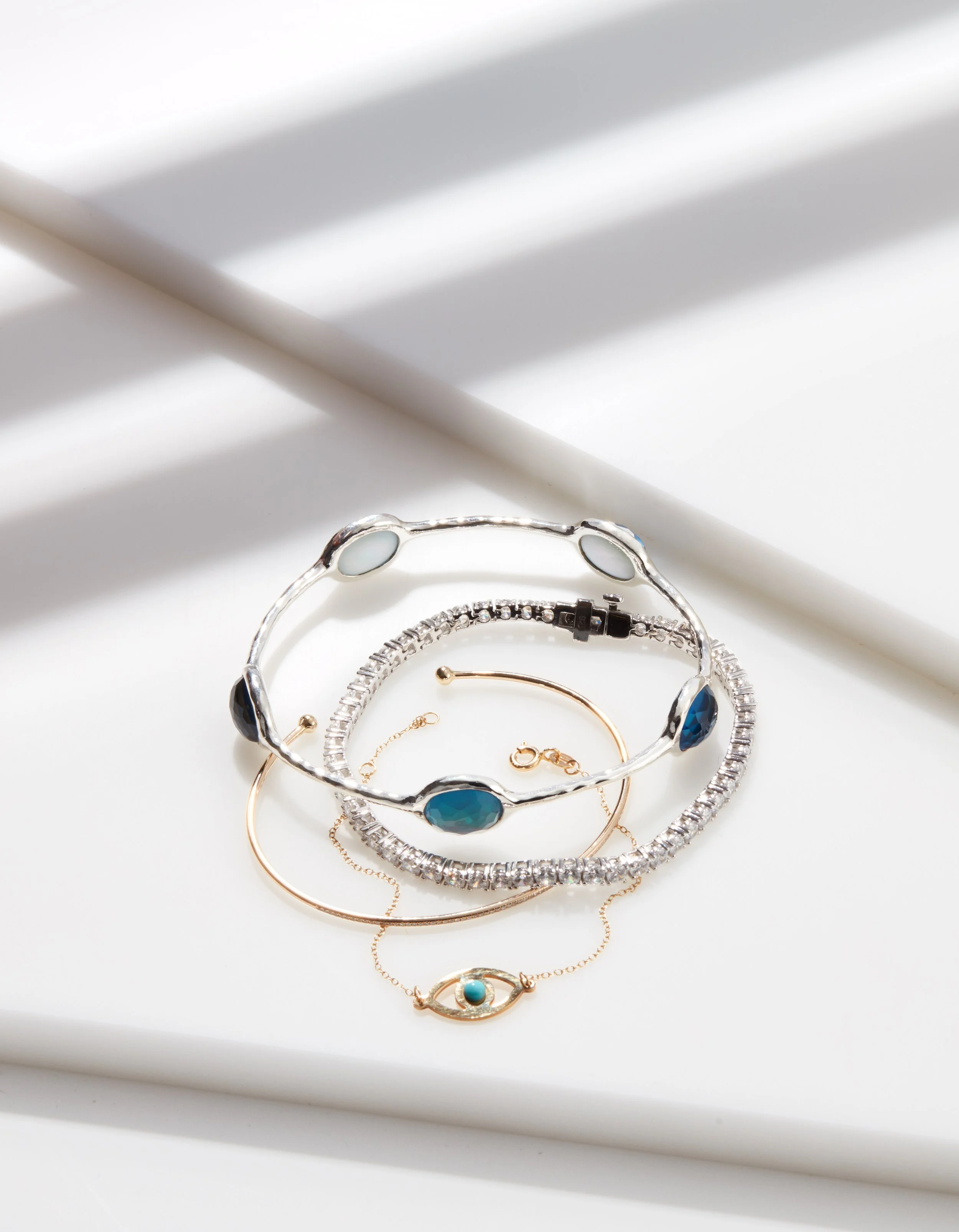 Collection of silver, gold, and chain bracelets with blue and clear gemstones displayed on a white surface with sunlight shadows.