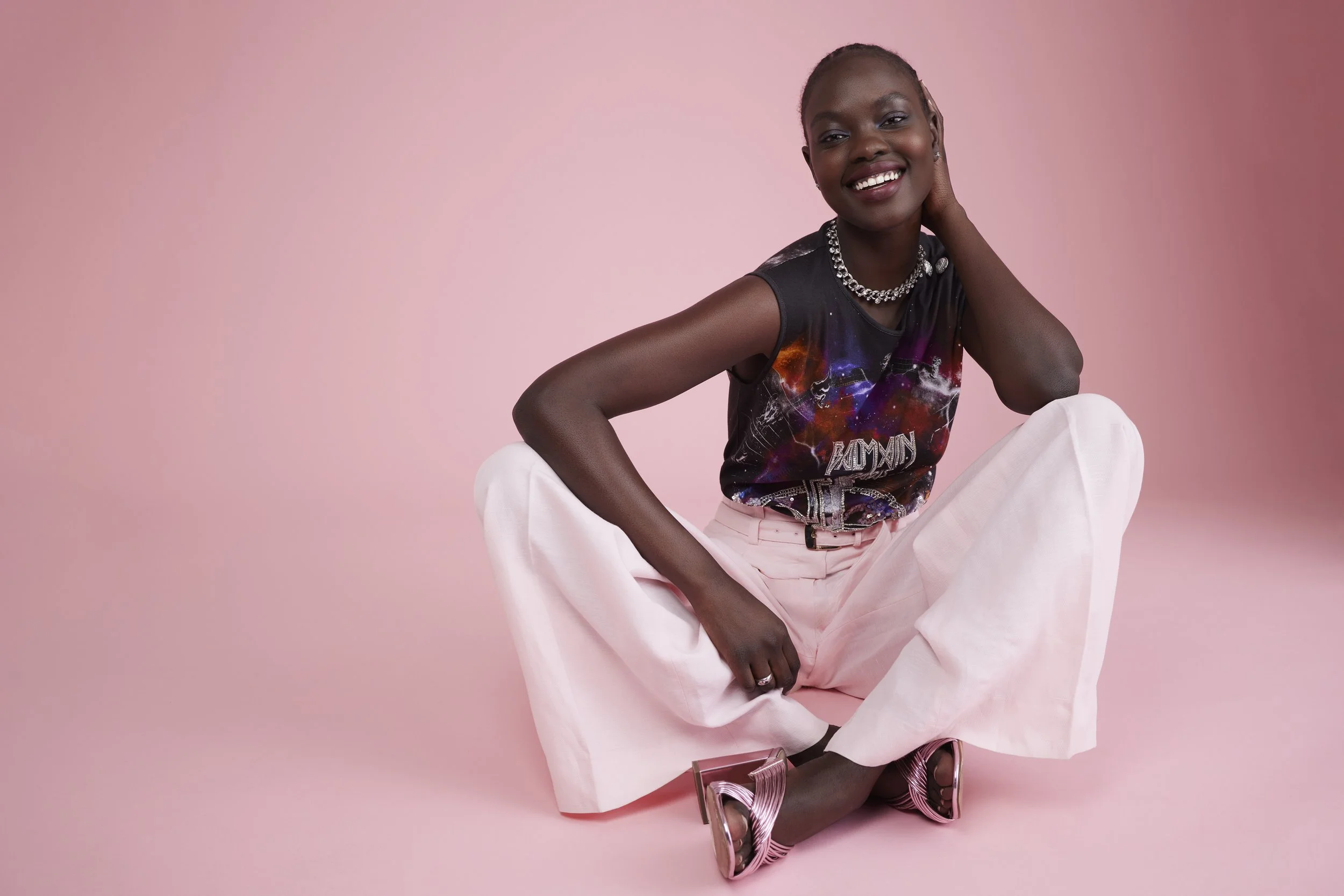 A woman sitting on the floor against a pink background, smiling and touching her neck, wearing a galaxy-themed sleeveless top, pink pants, metallic high-heeled shoes, and a chunky silver necklace.