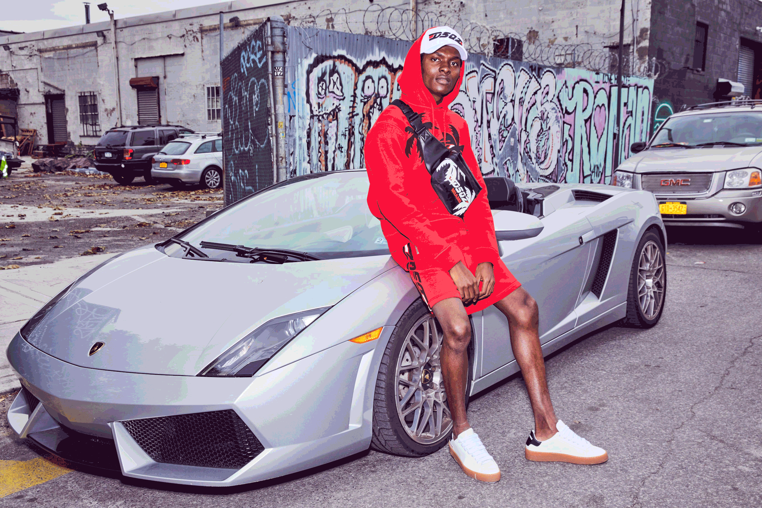 A young man dressed in red sportswear leaning against a silver Lamborghini in an urban lot with graffiti walls and parked cars in the background.