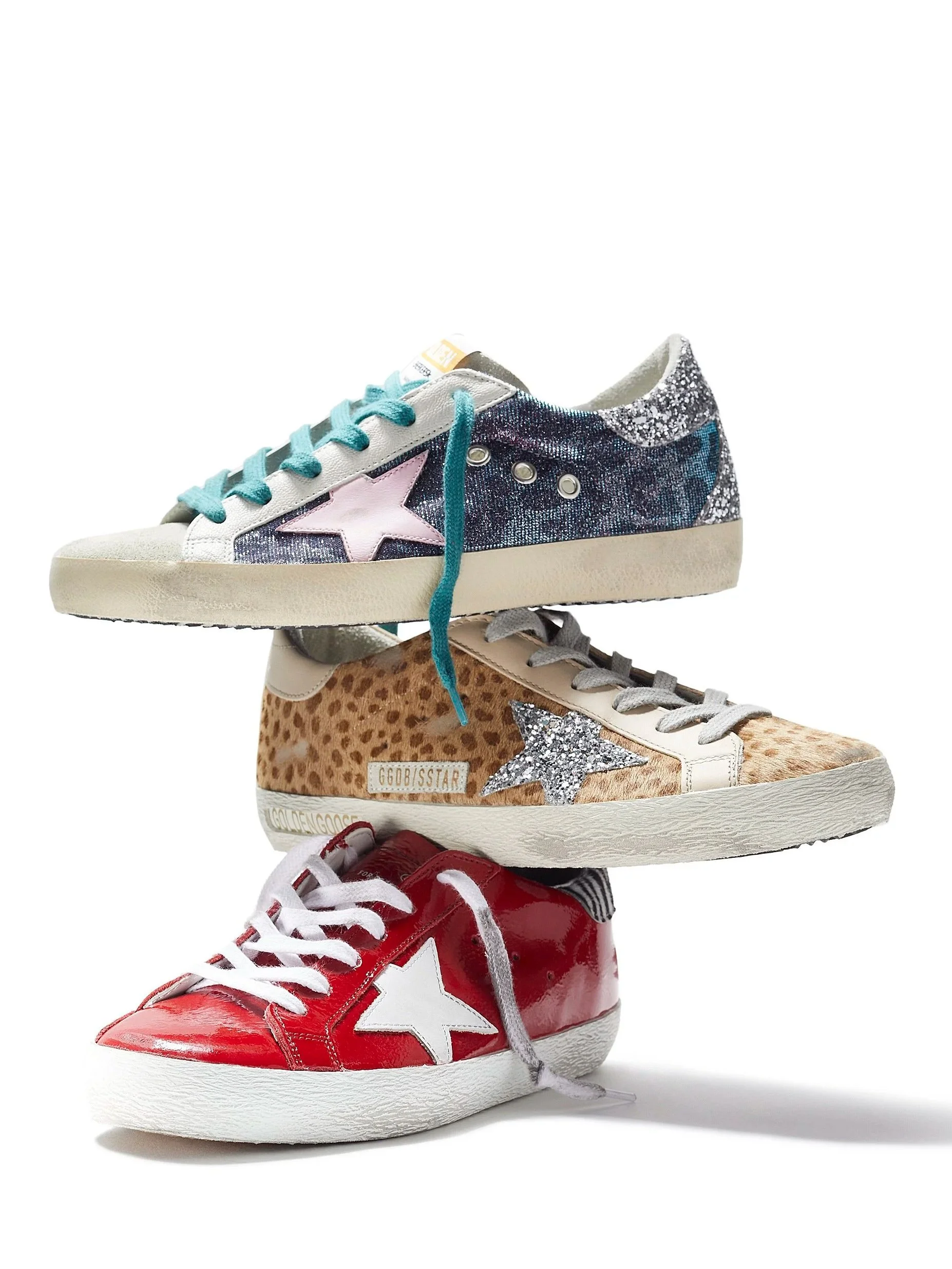 Multiple sneakers stacked vertically, including a red and white sneaker at the bottom, a beige and leopard print sneaker in the middle, and a glittery dark blue sneaker with a star and pink star details on top.