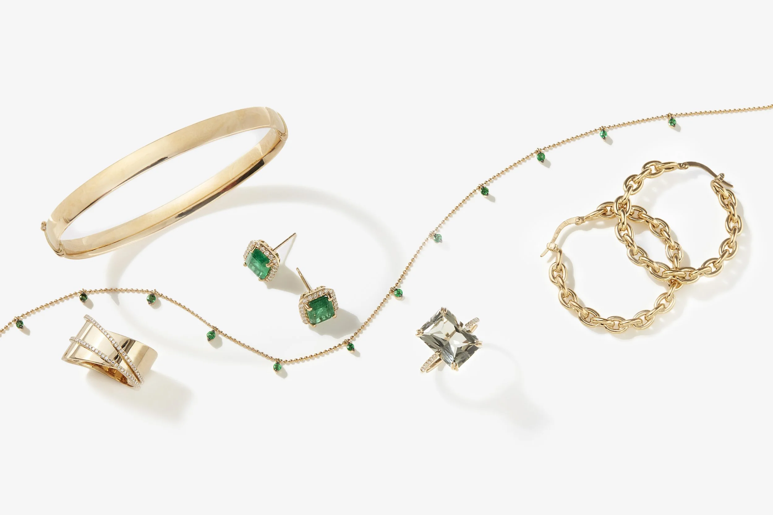 Assorted gold jewelry including bracelets, necklaces, rings, and earrings, some featuring green gemstones, arranged on a white background.
