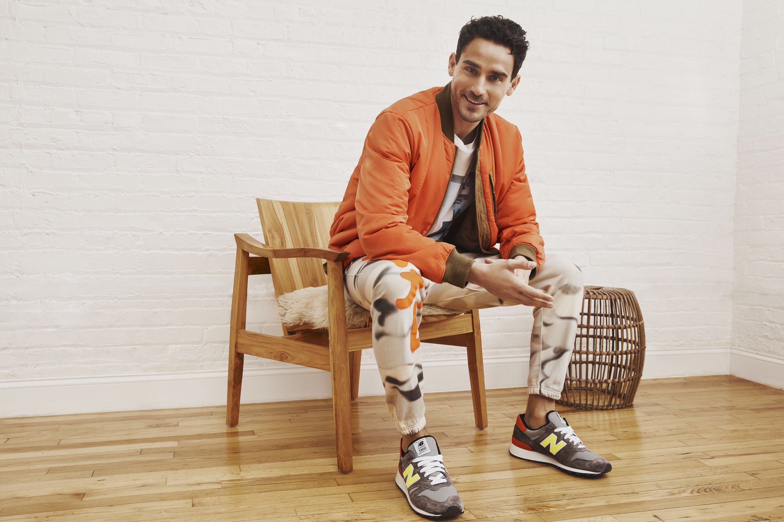 A young man sitting on a wooden chair in a room with a white brick wall. He is wearing an orange jacket, patterned sweatpants, and sneakers, smiling and looking slightly to the side.