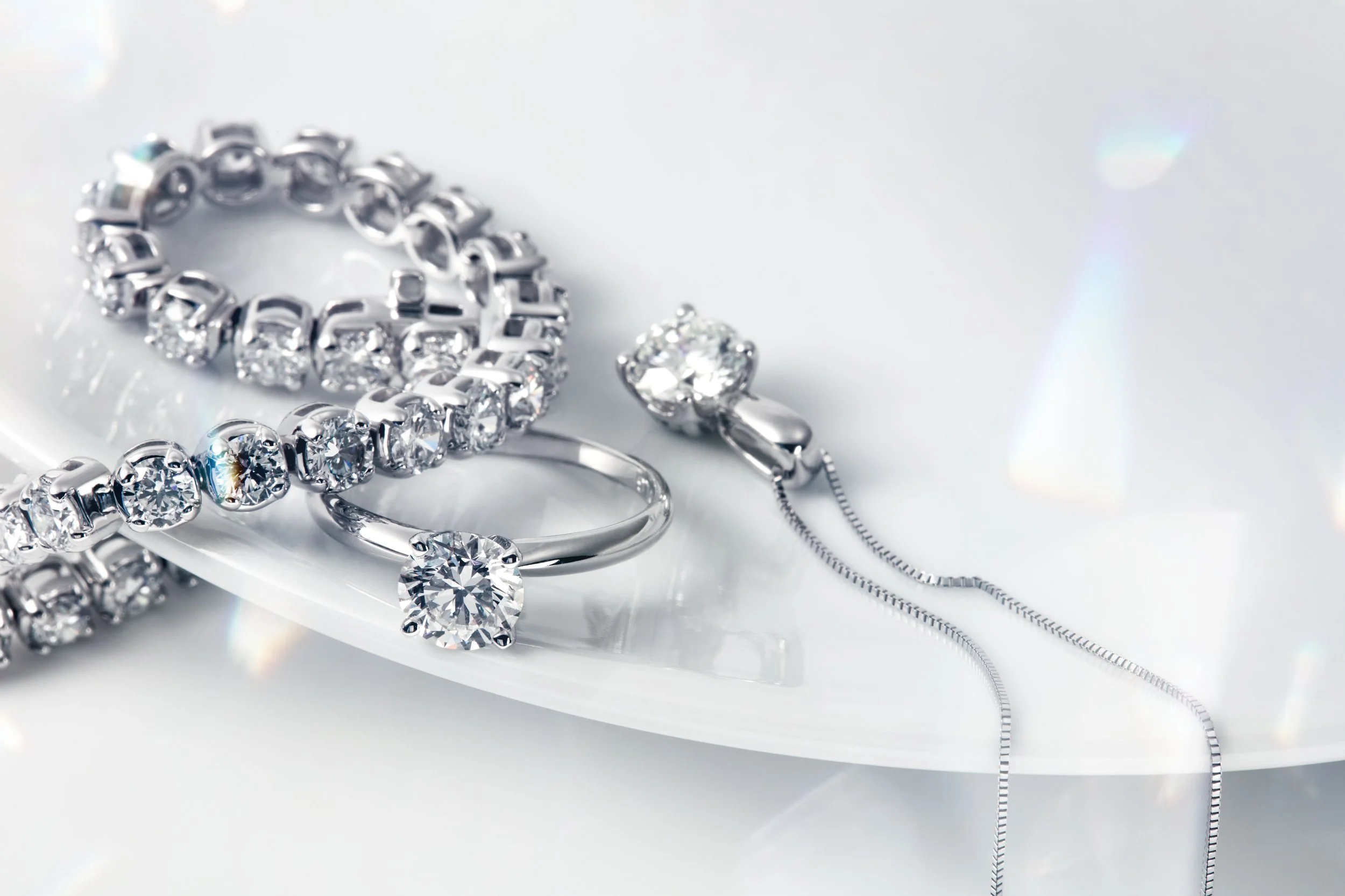 Collection of silver jewelry with sparkling gemstones, including a ring, bracelet, necklace, and earrings, arranged on a white reflective surface.