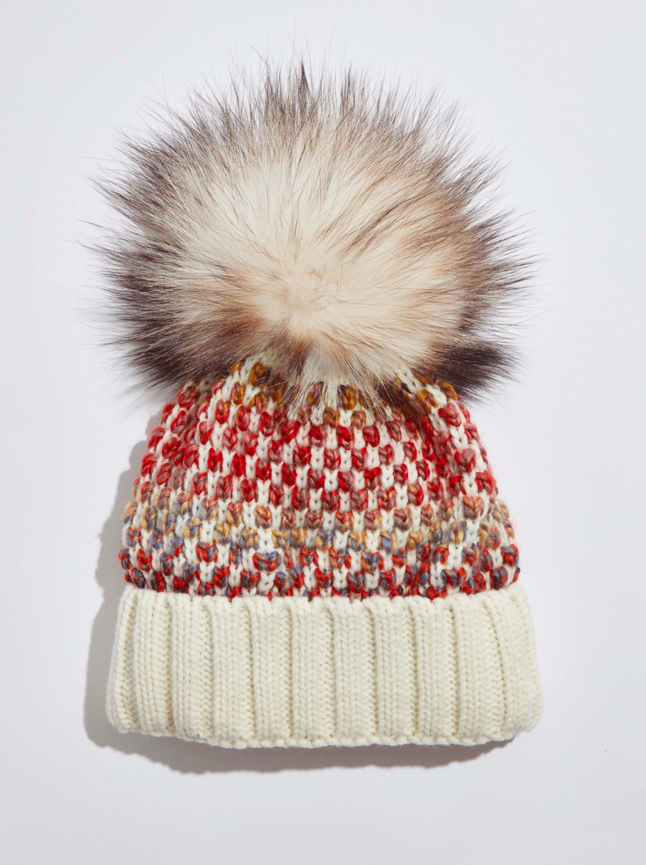 A close-up of a knitted winter beanie hat with a large faux fur pom-pom on top, against a plain background.